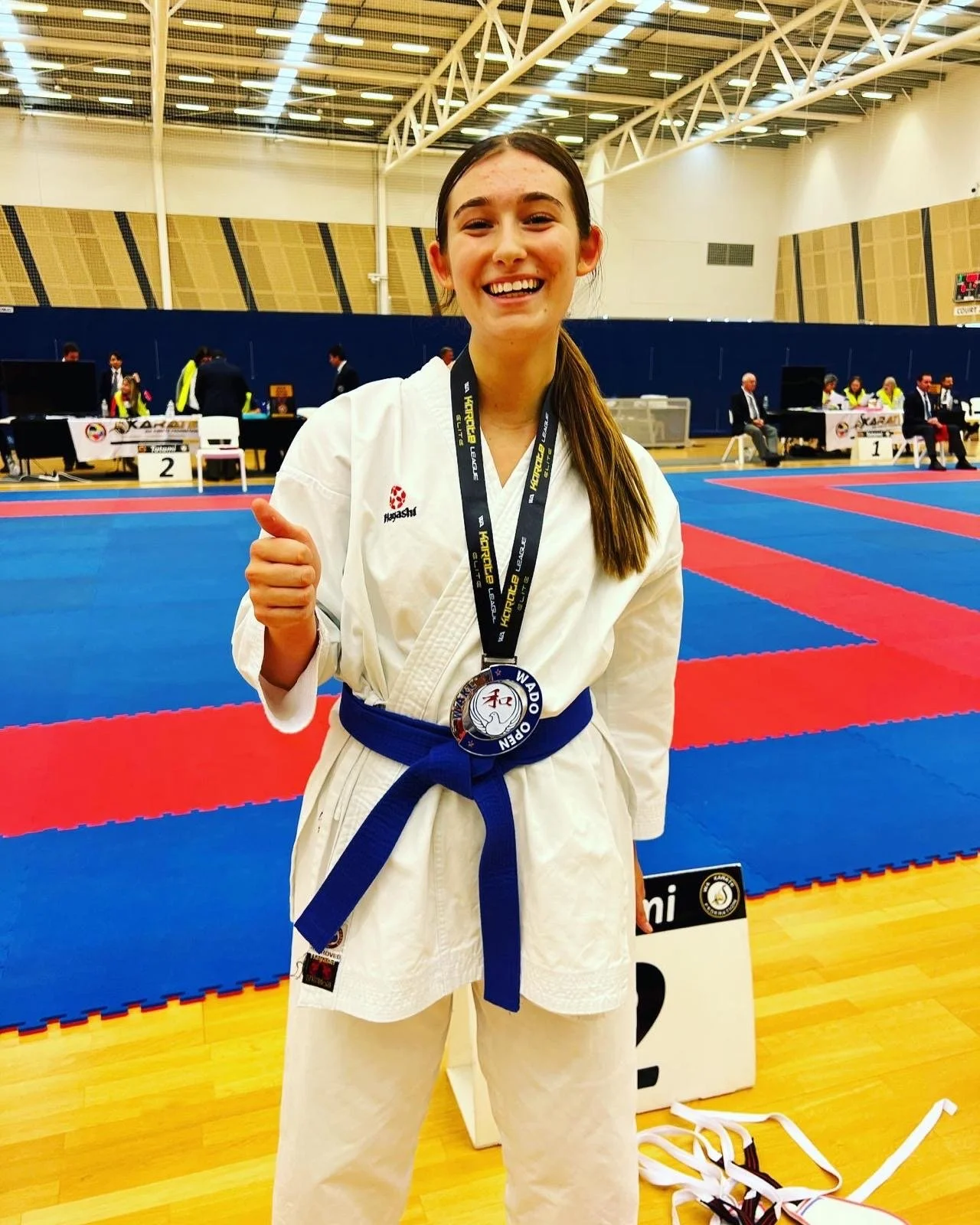 A young female karate athlete in a white gi with a blue belt, smiling and giving a thumbs-up, standing on a competition mat inside a sports hall. She has a medal around her neck, and there are judges and officials in the background.
