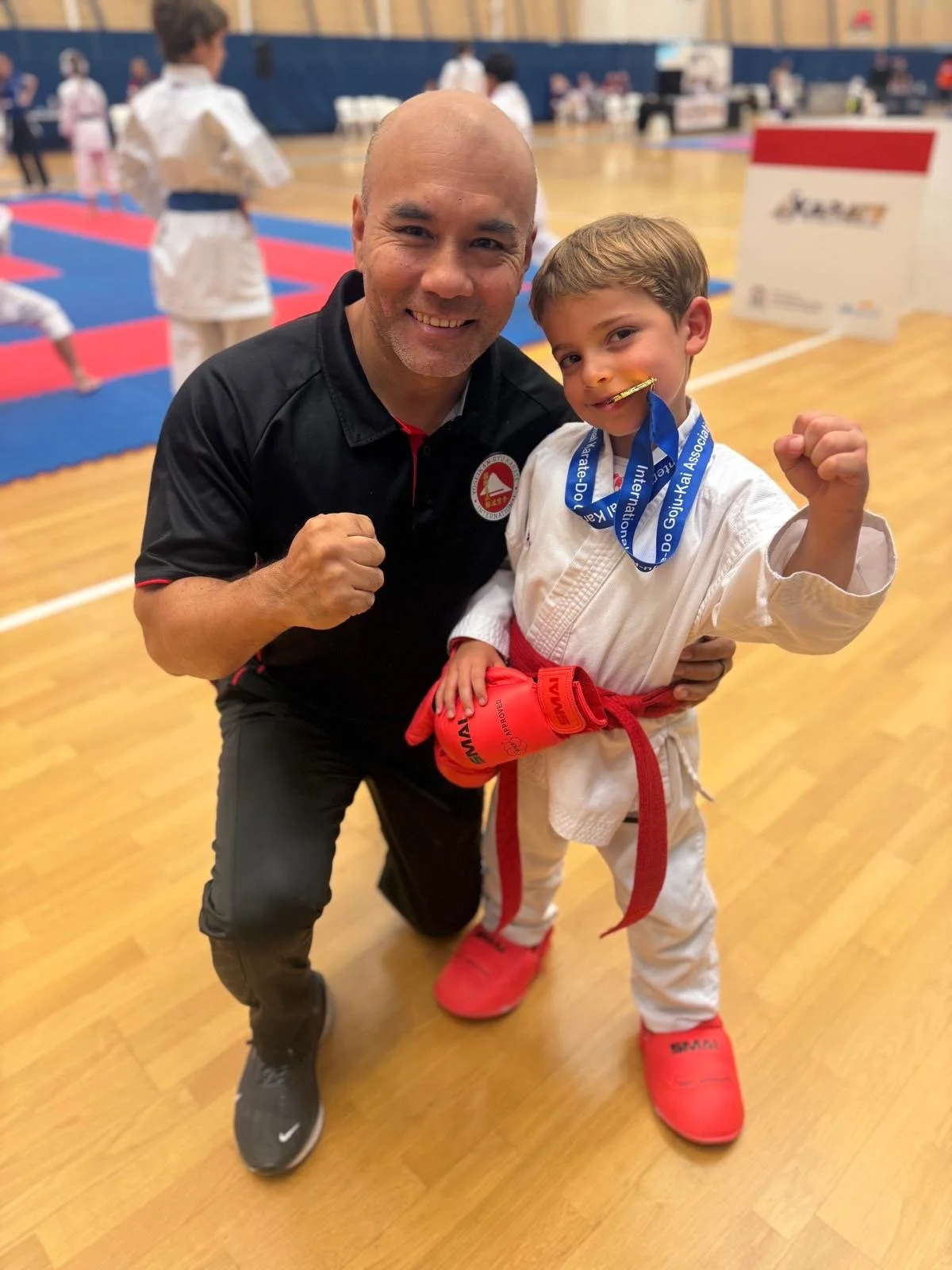 A young boy in a karate gi with a red belt and red gloves holding a medal, posing with a coach or instructor in a karate dojo or gym, both smiling, with other children practicing karate in the background.