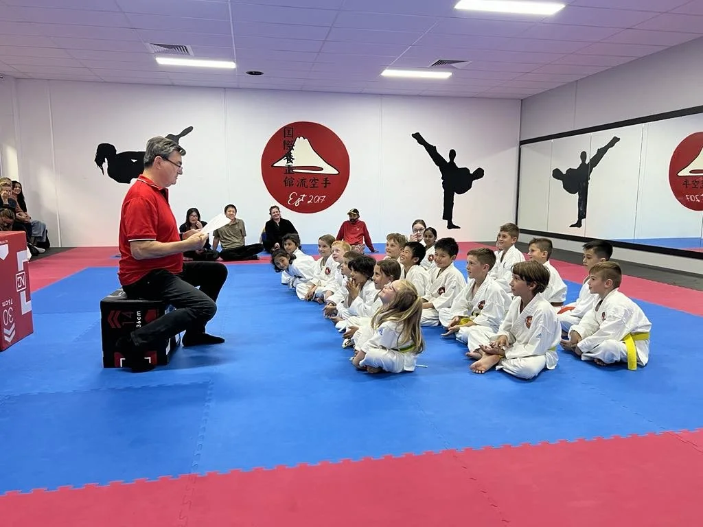 A martial arts instructor in a red shirt sitting on a black box, reading from a paper to a group of children in white gi uniforms with yellow belts, seated on a blue and red mat in a martial arts dojo. The walls display gymnastic silhouettes and a re