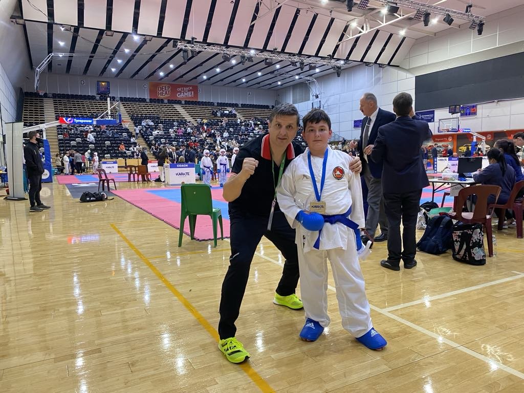 A young martial artist in a white karate gi with a blue belt and gloves standing next to an adult man in a black jacket and bright yellow shoes inside a sports arena. The arena has a martial arts competition setup with mats, judges, and spectators in