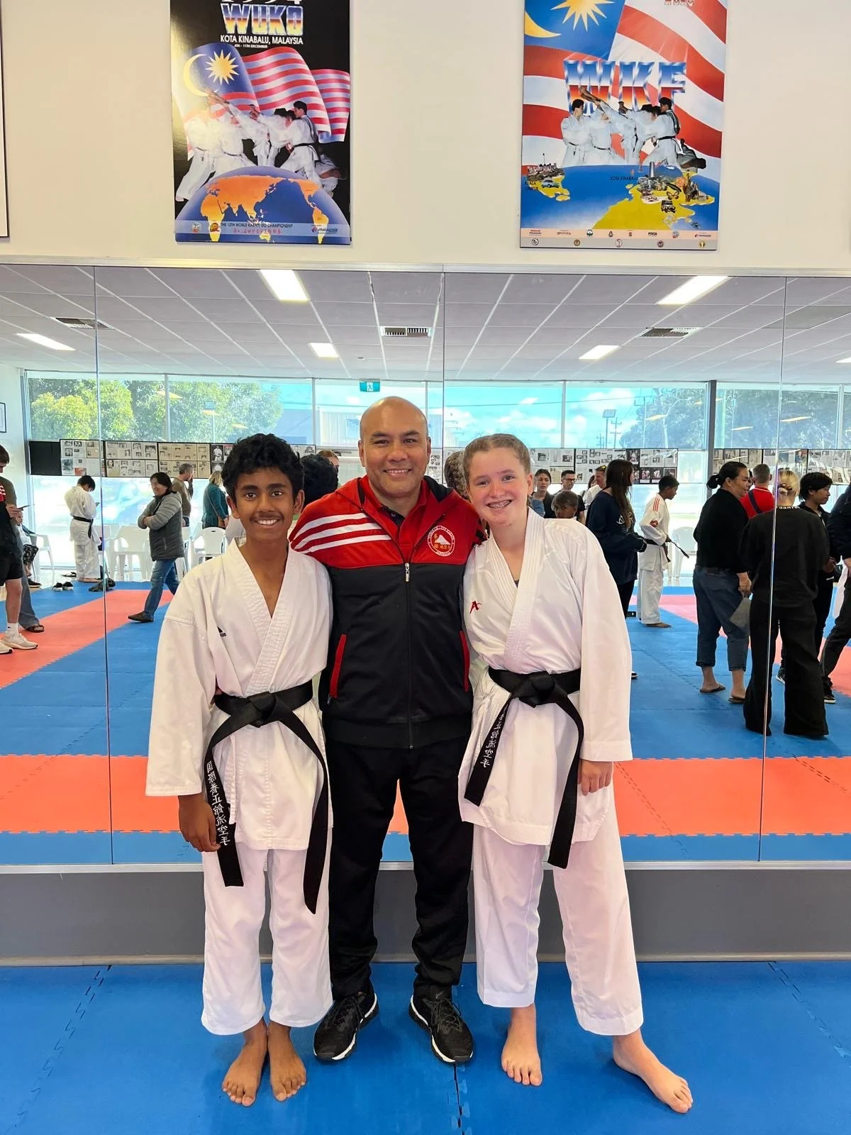 Two young female martial artists in white gi with black belts standing with a male coach in a martial arts dojo. The dojo has mats and other students training in the background, with large posters of martial arts on the wall.