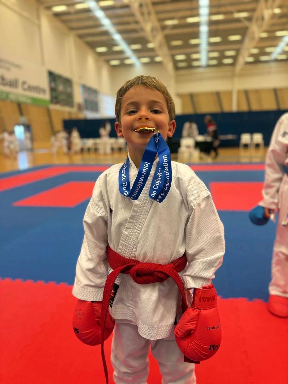 Young boy dressed in a judo gi with a red belt and red boxing gloves, smiling and holding a gold medal with a blue ribbon in his mouth, standing on a red and blue martial arts mat in a gymnasium.