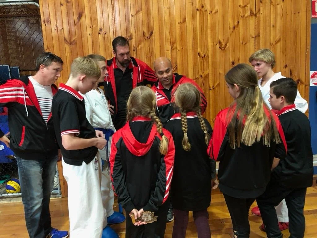 Group of young athletes in black and red tracksuits listening to coach in indoor sports facility.