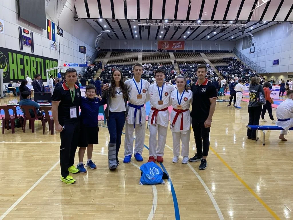 Group of seven people, including two children, standing on a gymnasium floor, some wearing medals and martial arts uniforms, at a sports event.