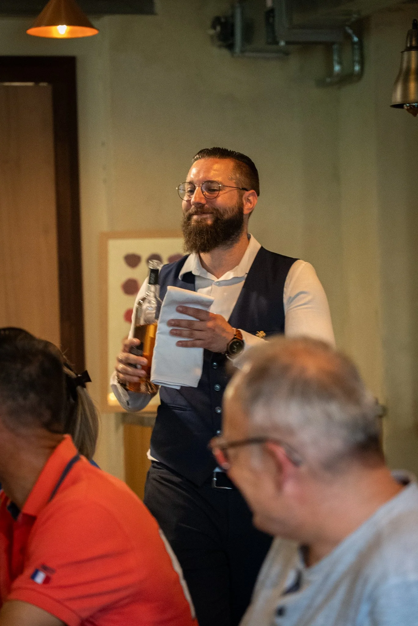 Un homme barbu, portant des lunettes, une chemise blanche et un gilet noir, tient une bouteille de bière et un torchon dans un restaurant ou un pub. Il apparaît à l'air souriant ou satisfait, entouré de personnes assises à une table.