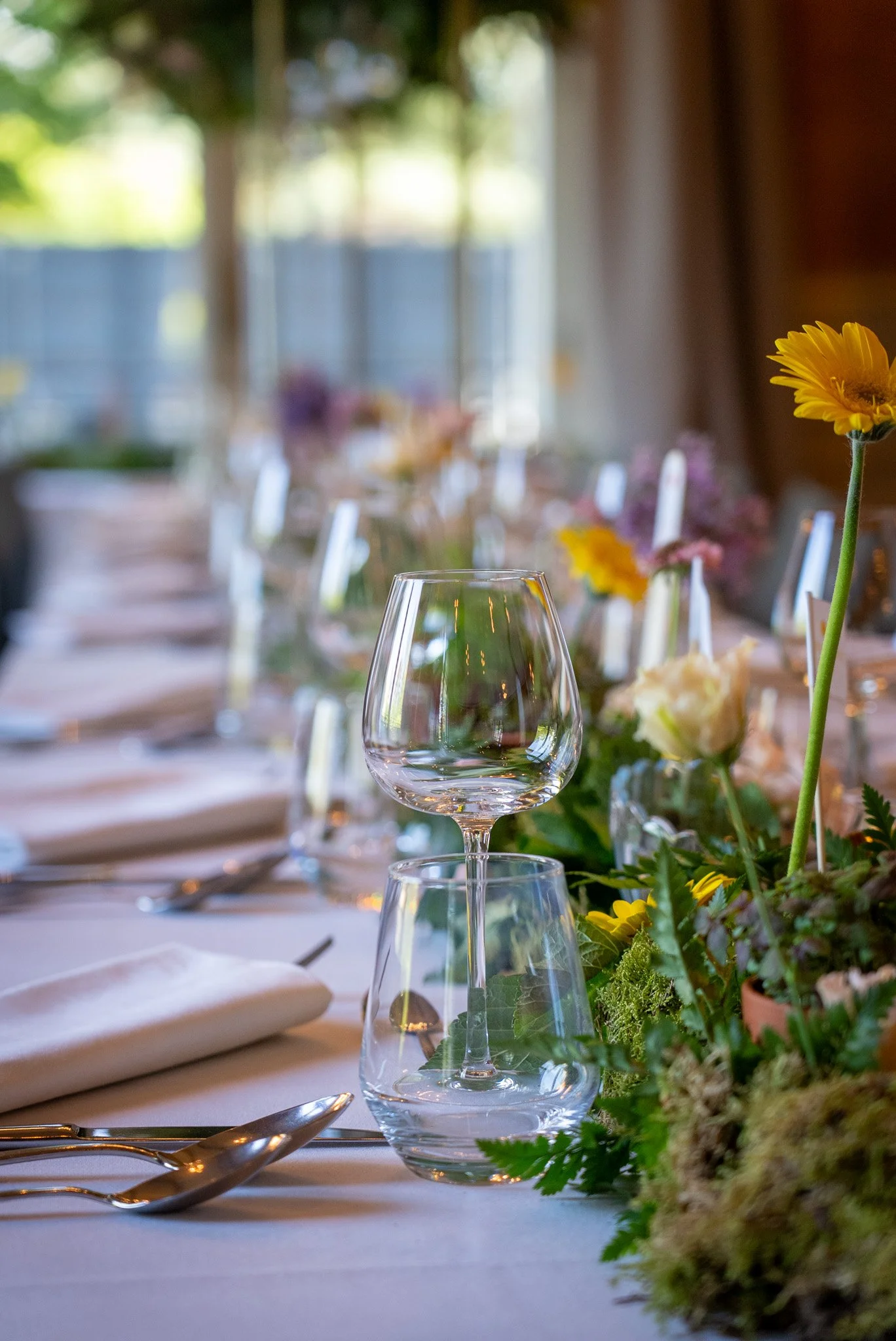 Table dressée pour un repas avec un centre de fleurs, des verres à vin et des assiettes, dans une ambiance lumineuse et printanière.