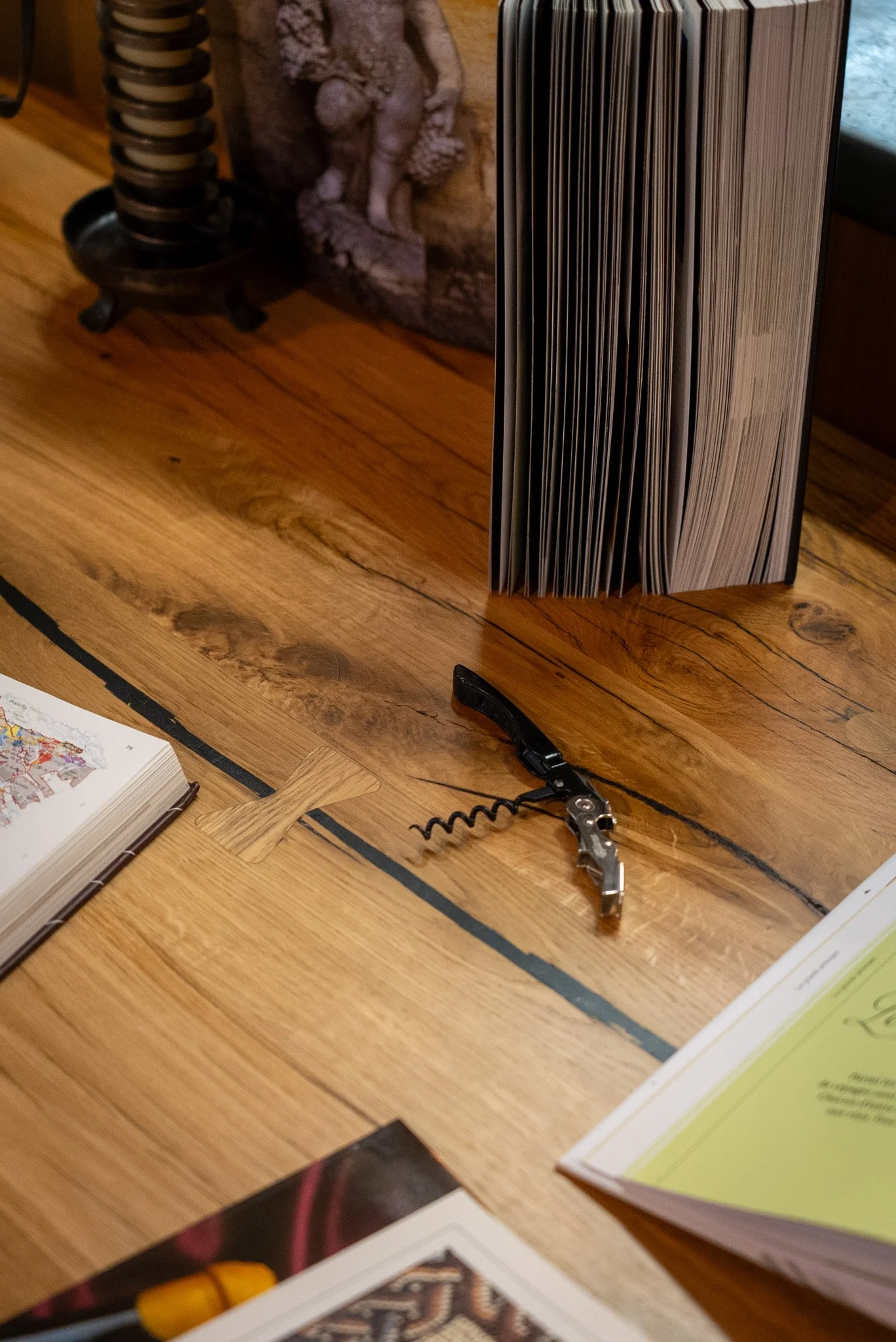 Objet sur une table en bois, comprenant un livre fermé, un ouvre-bouteille à tire-bouchon, et quelques documents ou livres.