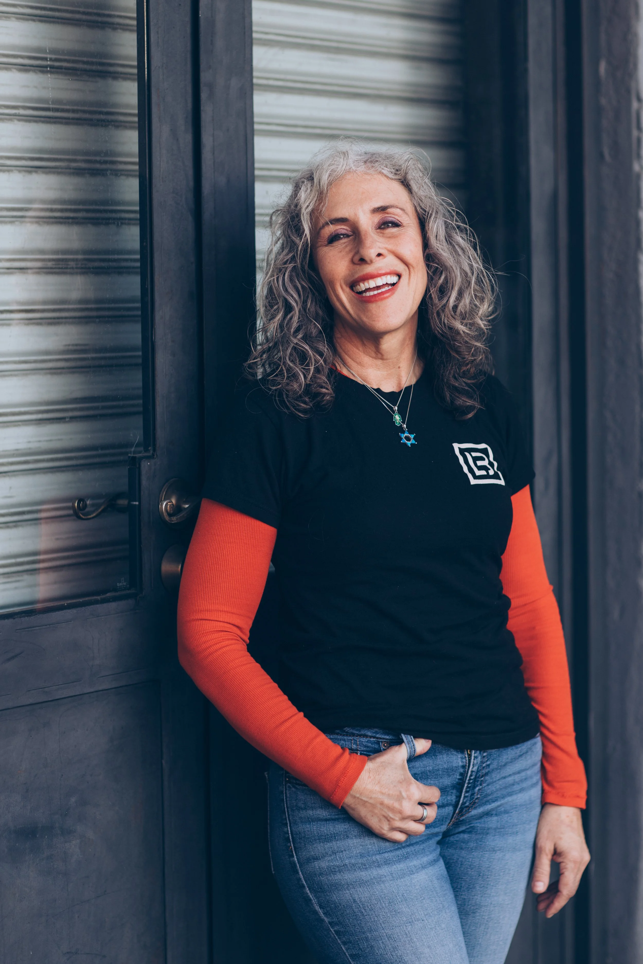 Smiling person with curly gray hair wearing a black T-shirt and jeans, standing against a gray door.