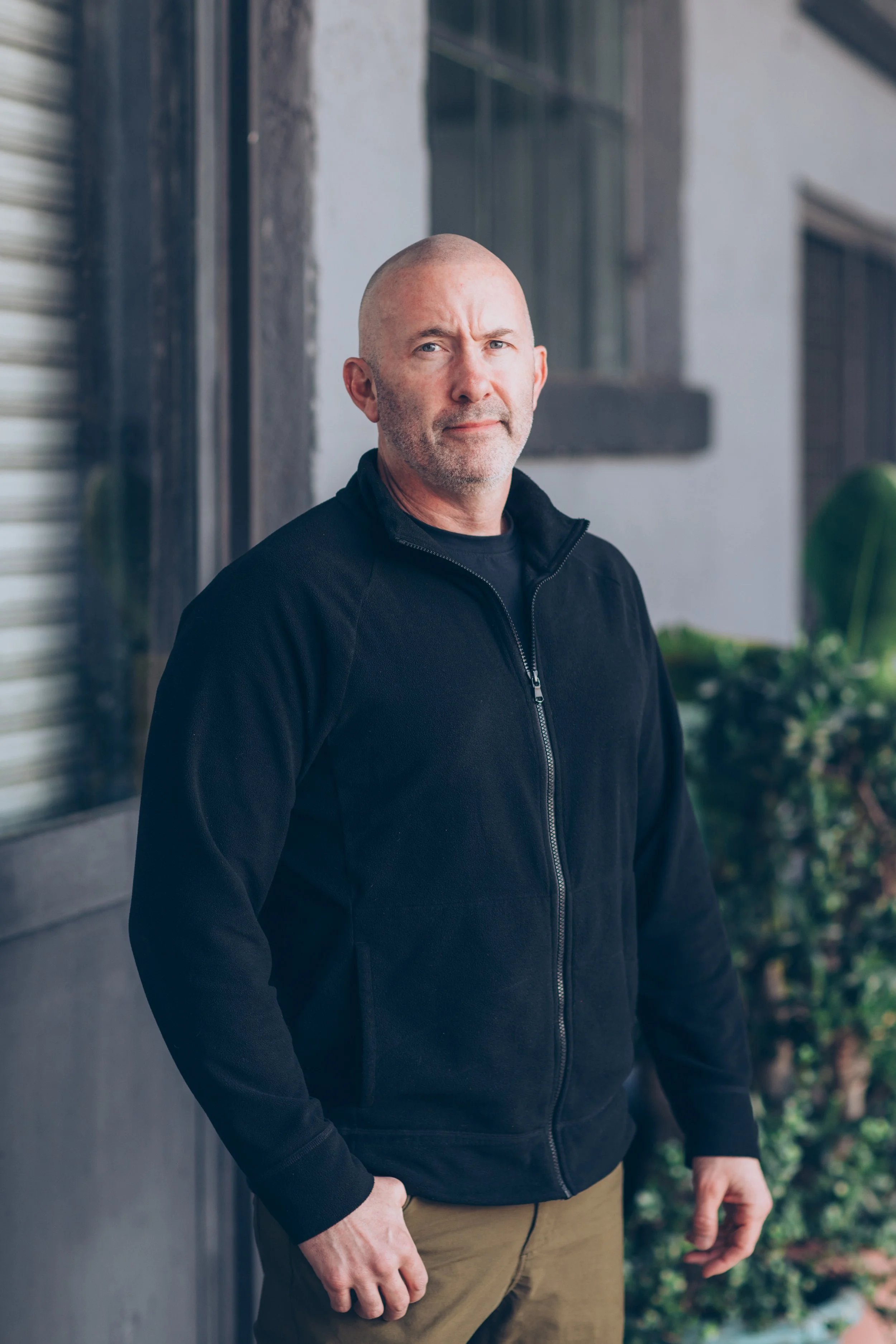 Man in a black jacket standing outdoors next to a wall with greenery.