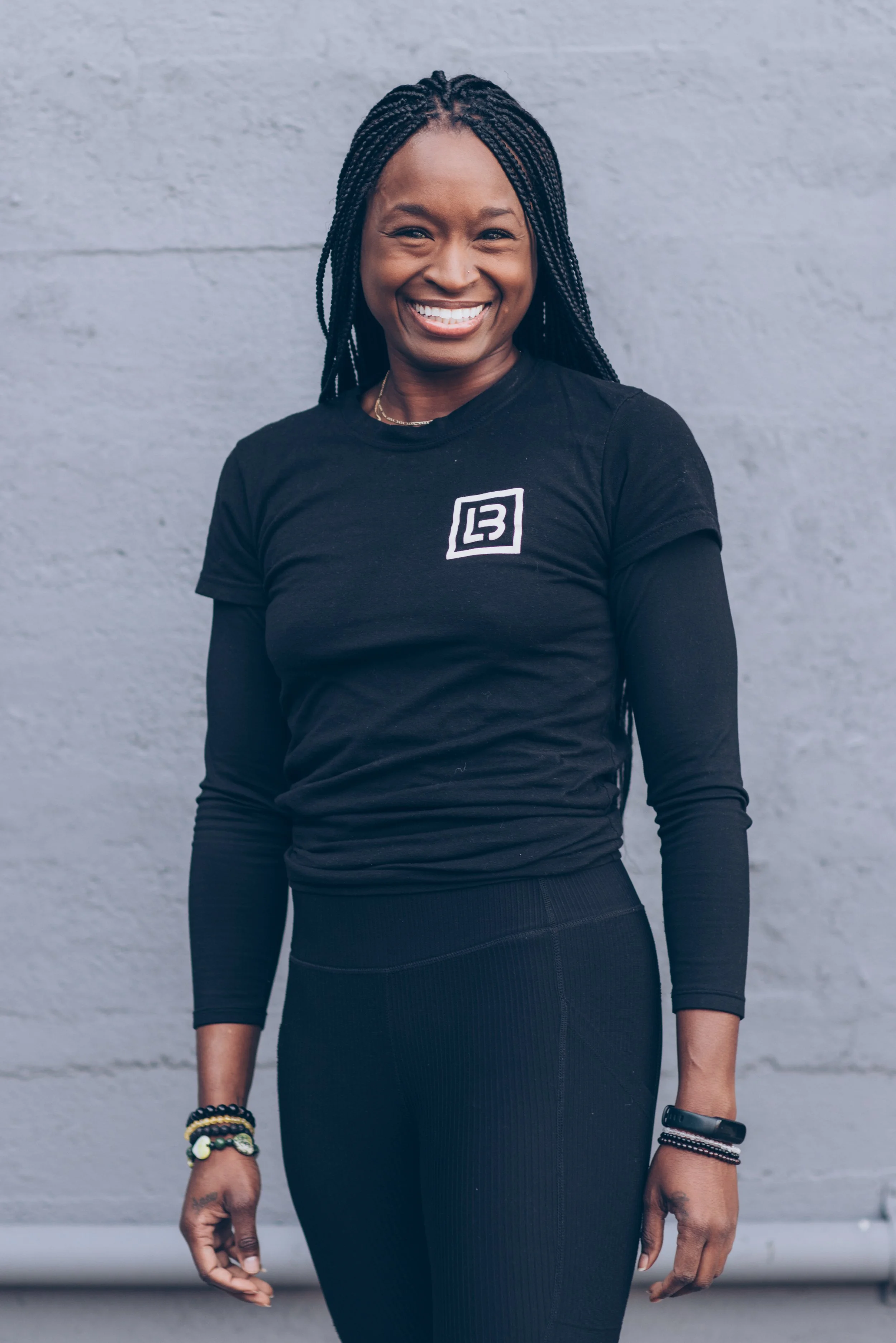 Smiling person with braided hair wearing a black long-sleeve shirt and bracelets against a gray background.