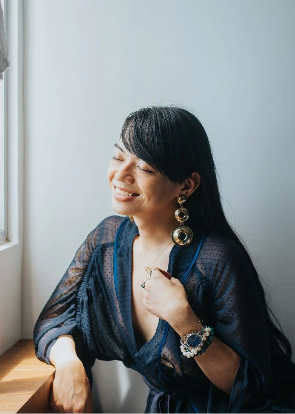 A woman with black hair, wearing large gold earrings, a watch, and a navy blue sheer blouse, sitting by a window, smiling and enjoying a peaceful moment.