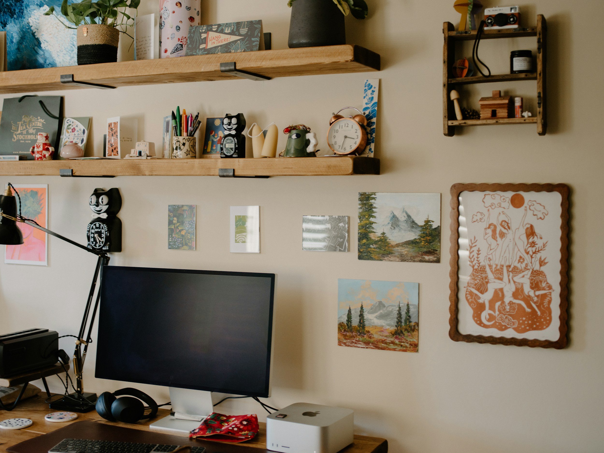 A picture of a black laptop on a brown desk with pictures of artwork on the walls. A cat-shaped clock and wall shelves.