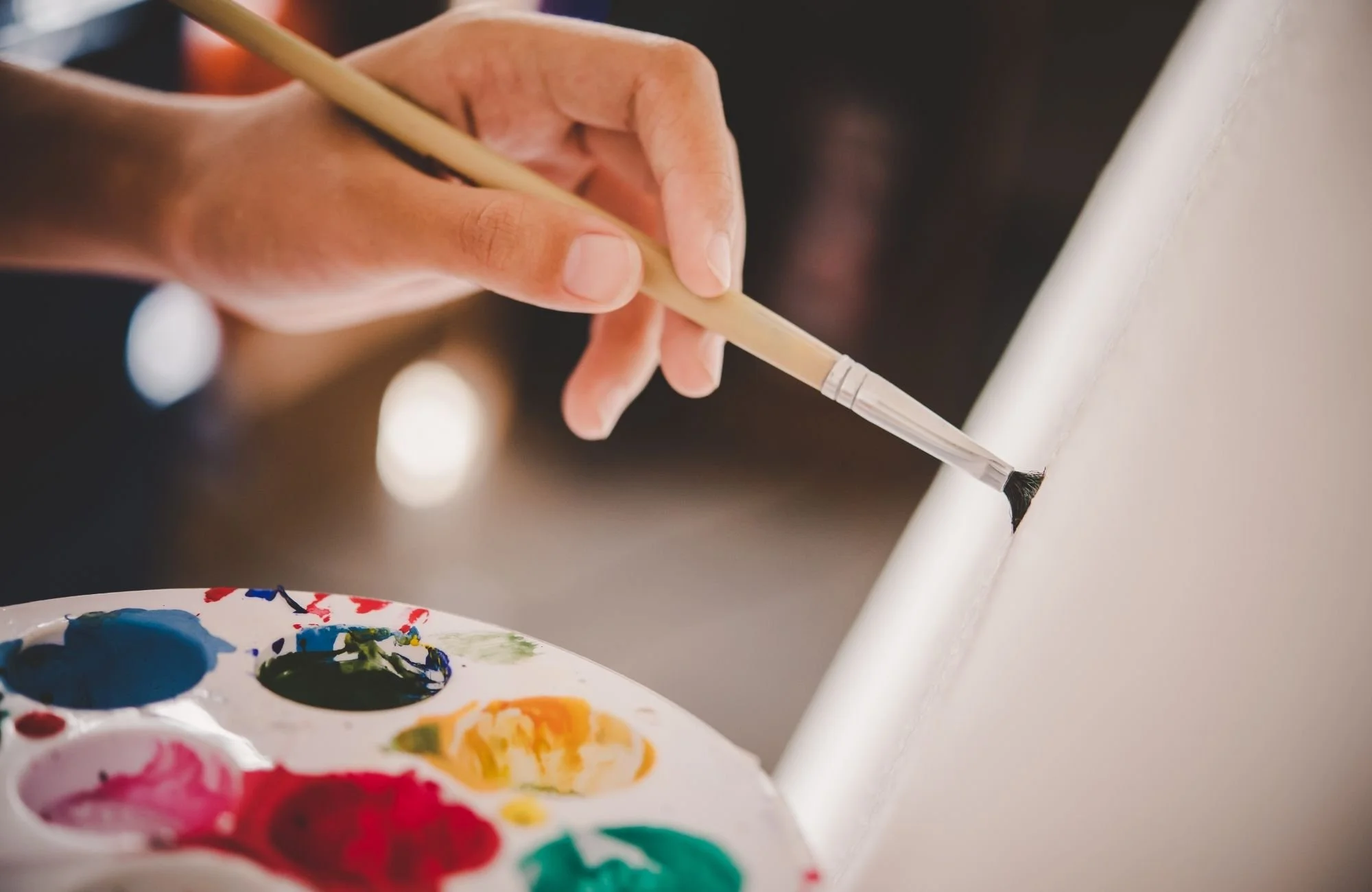 Close-up of a person using a paintbrush to paint on a white canvas, with a palette of colorful paints nearby.