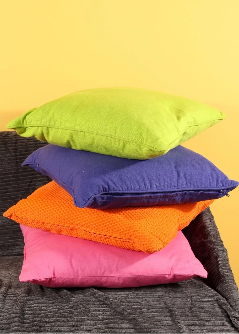 A stack of five colorful pillows on a dark gray textured couch against a yellow wall.