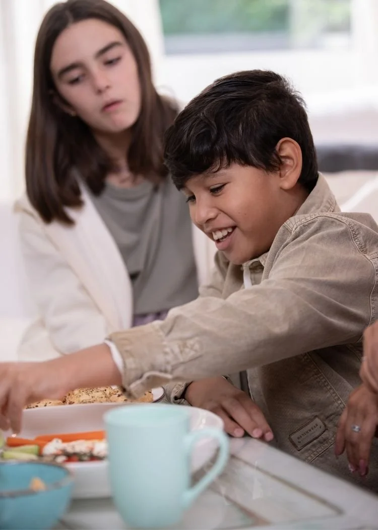 A young boy smiling and reaching for food on a plate at a kitchen counter, with a girl standing beside him watching.
