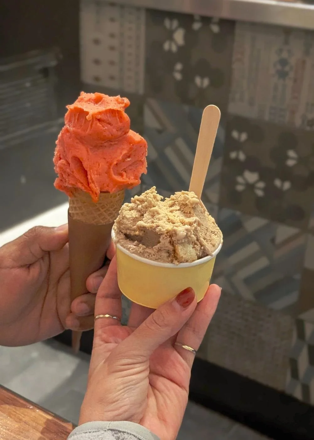 Two people holding ice cream cones, one with orange sorbet and the other with brown coffee or caramel flavor, at a store or cafe.