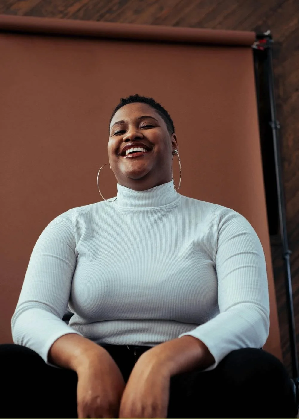 A black woman with short hair who's smiling with a white turtleneck and earrings.