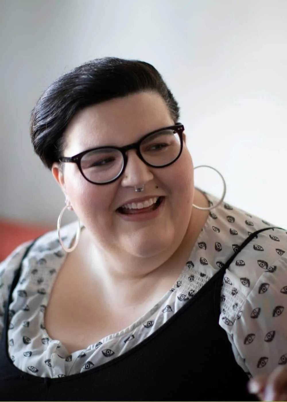 A smiling woman with short black hair, glasses, large hoop earrings, and a nose piercing, wearing a patterned top with eye designs.