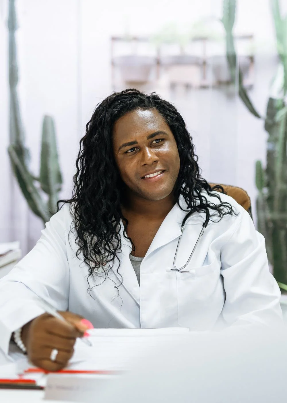 A male doctor with long, curly black hair sitting at a desk with a pen in hand, wearing a white coat and a stethoscope, smiling, with potted plants in the background.