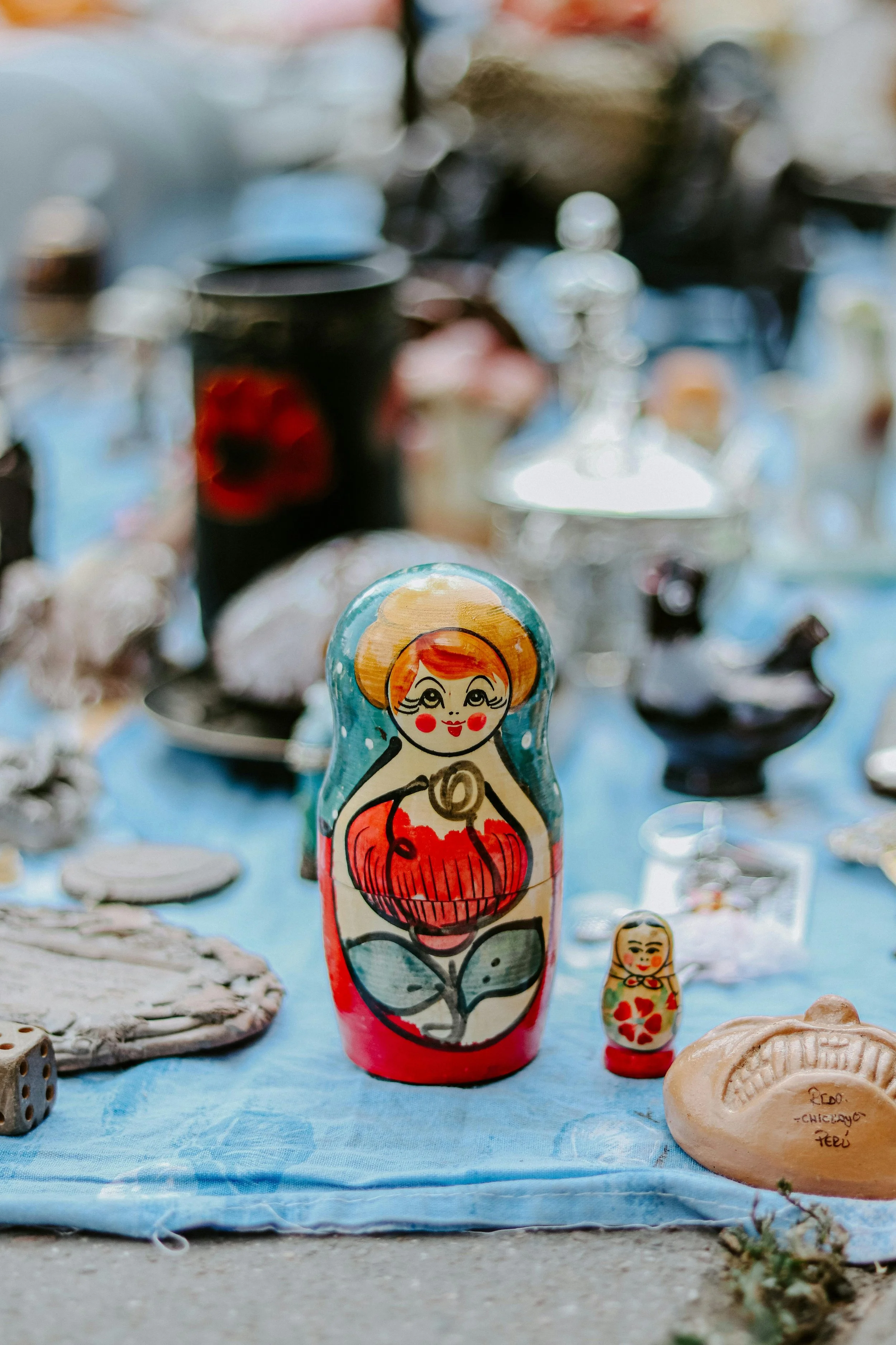 Colorful painted Russian nesting doll with a red dress and orange hair, on a blue cloth table with other decorative items in the background.