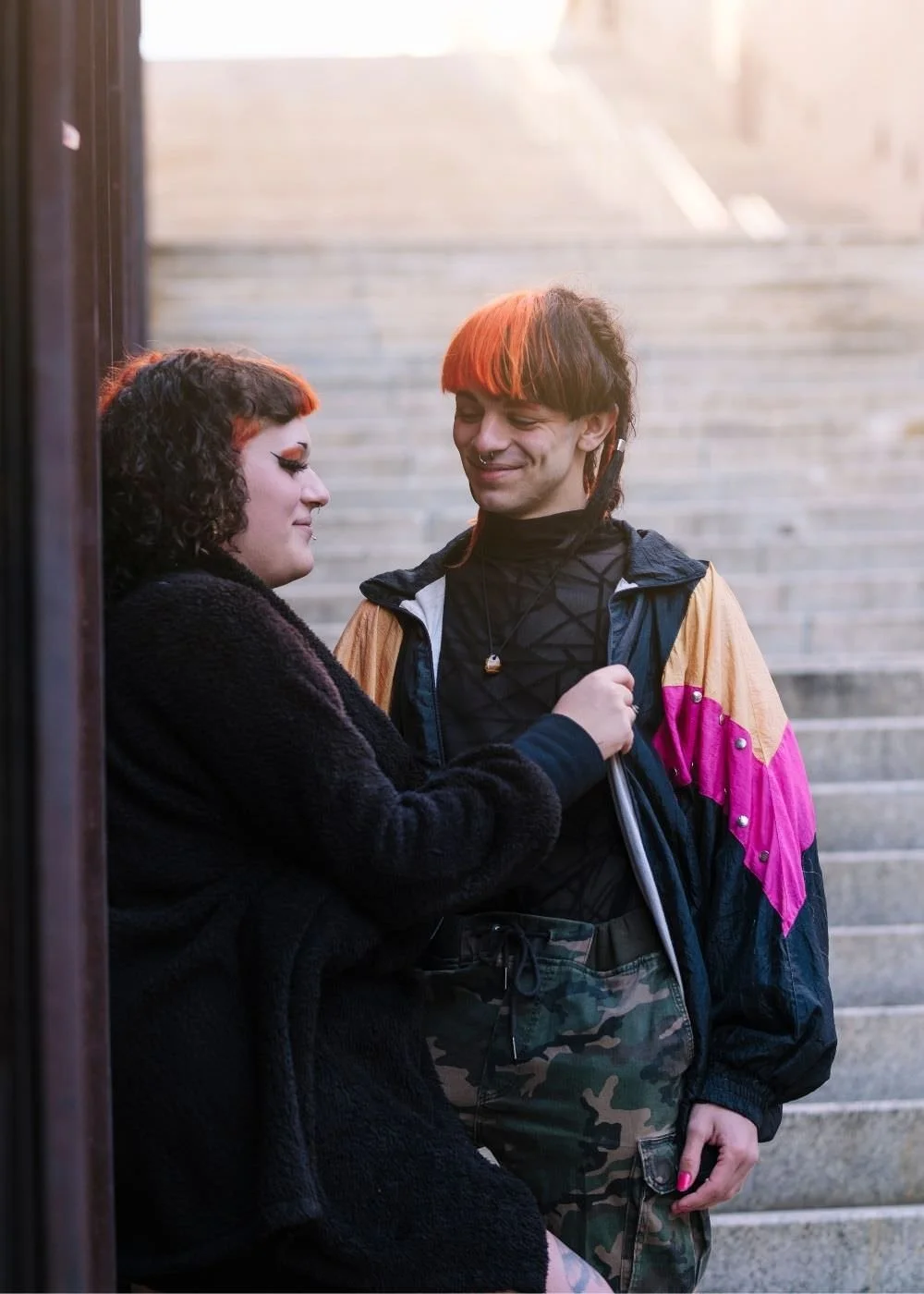 A picture of a couple with two transgender partners. One partner is leaning up against the wall while the other partner is smiling. Both have orange dyed hair.