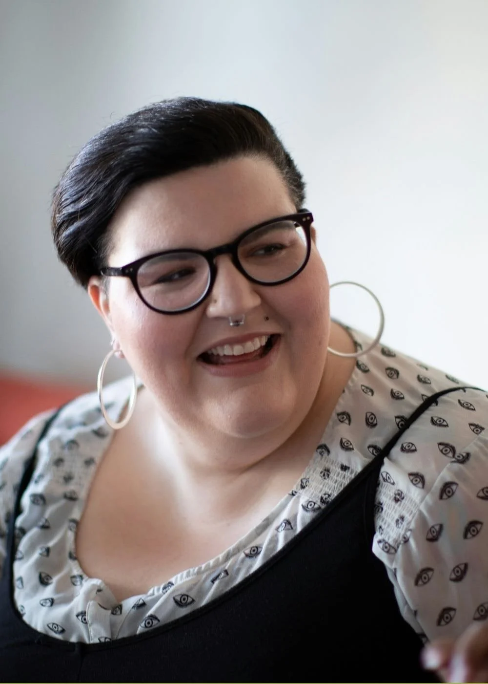 A picture of a non-binary person with glasses, short hair and who is smiling.