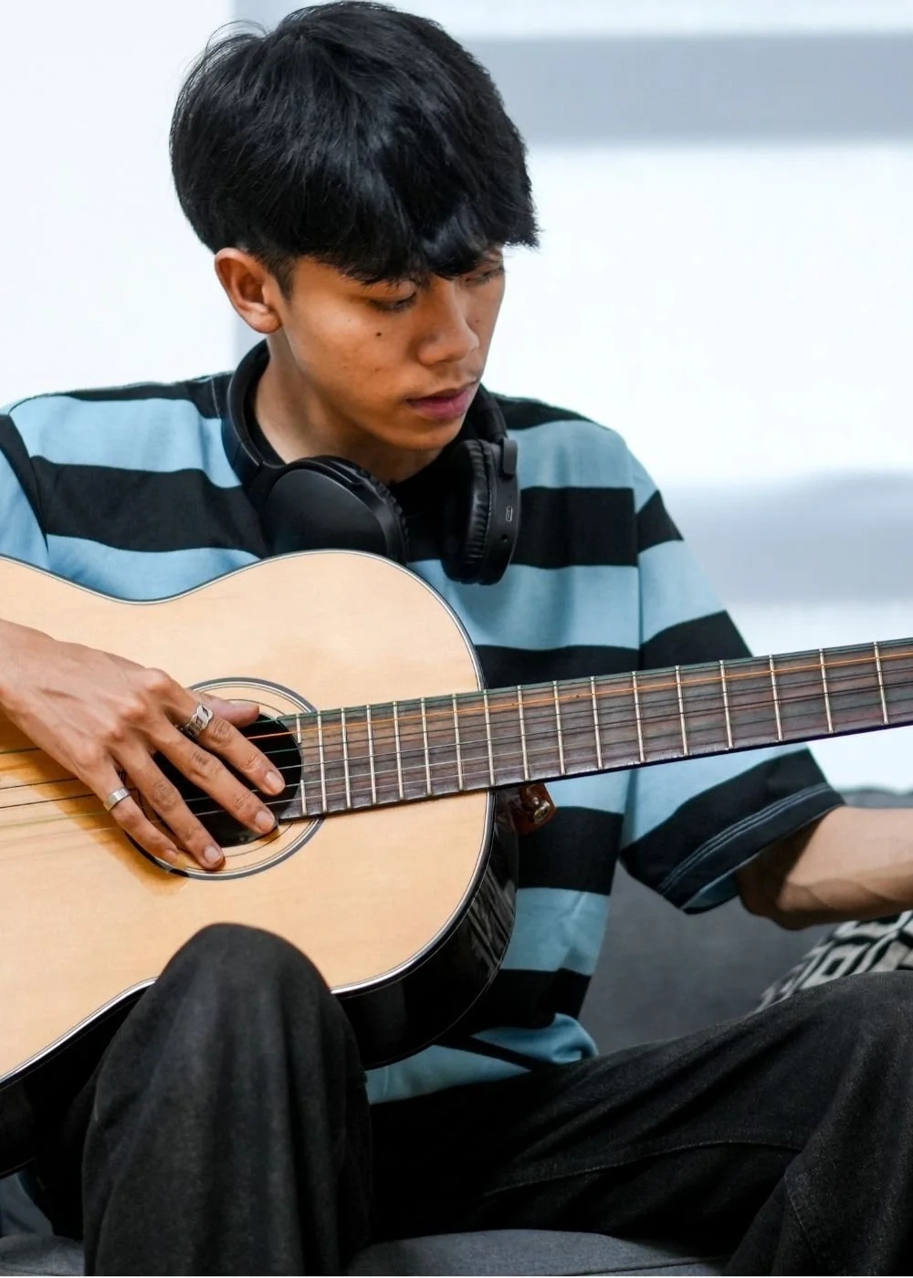 A young man with black hair wearing a striped shirt, sitting, playing an acoustic guitar with headphones around his neck, indoors.
