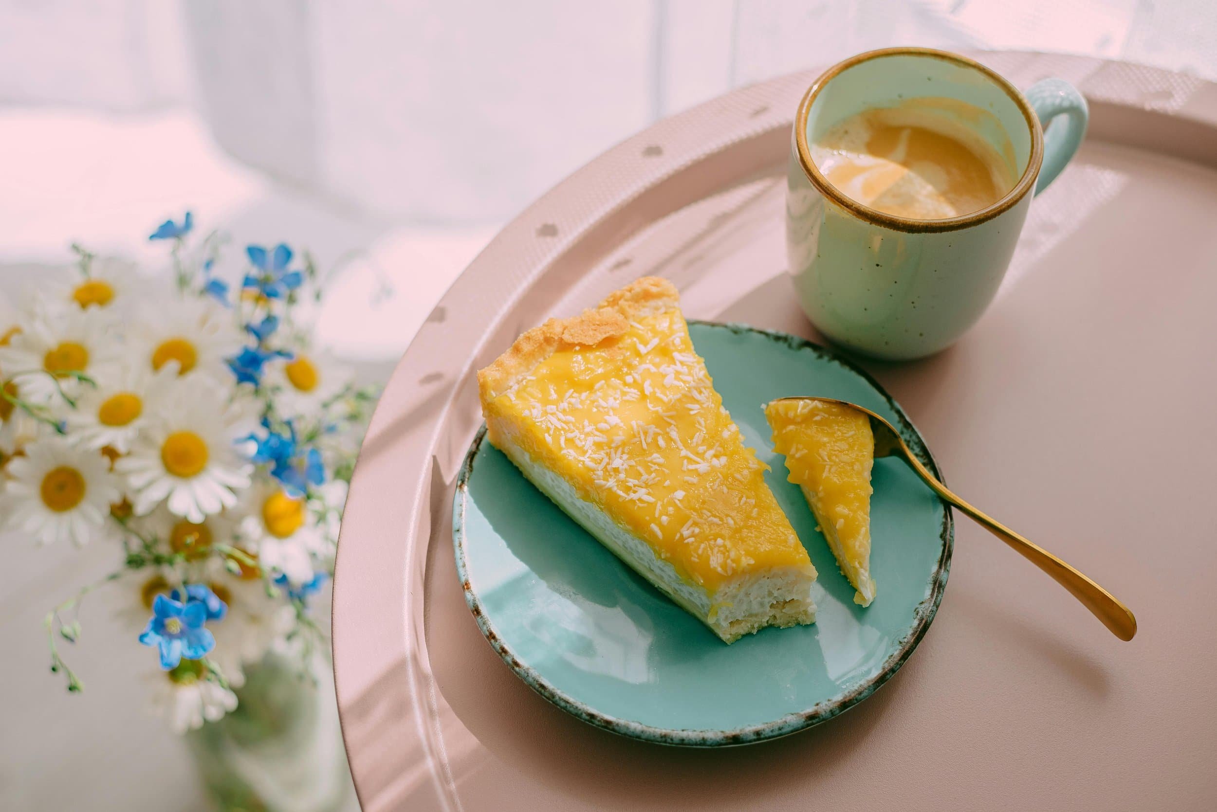 A slice of lemon cheesecake on a turquoise plate with a gold spoon, a mug of coffee, and a bouquet of daisies and blue flowers on a light-colored table.