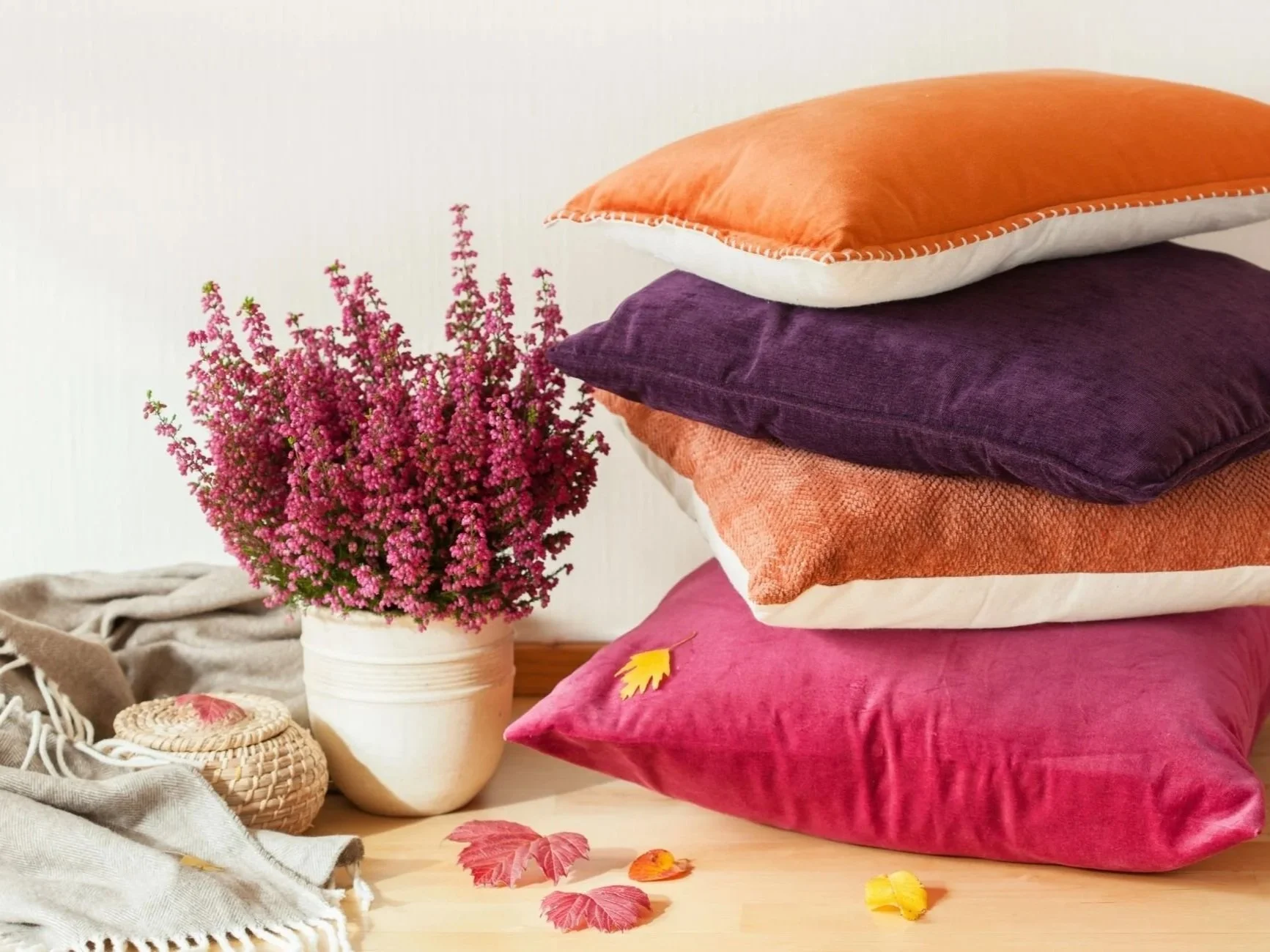 Stack of four colorful pillows in shades of orange, purple, peach, and pink on a wooden surface next to a white pot of pink flowers, with some fall leaves scattered on the surface.