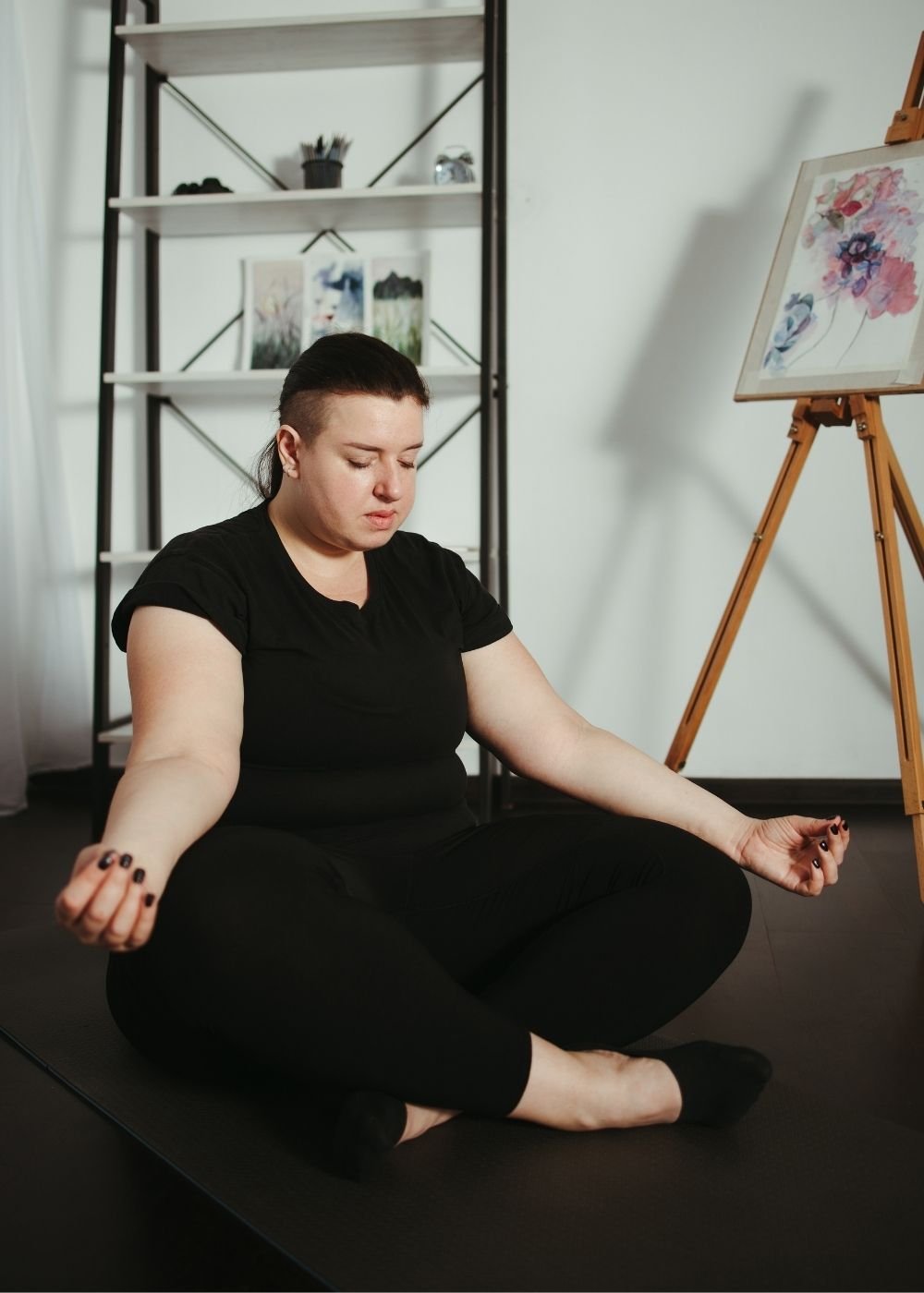 A gender non-conforming person practicing meditation while sitting cross-legged on a yoga mat in a room with an art easel and shelves with framed pictures.