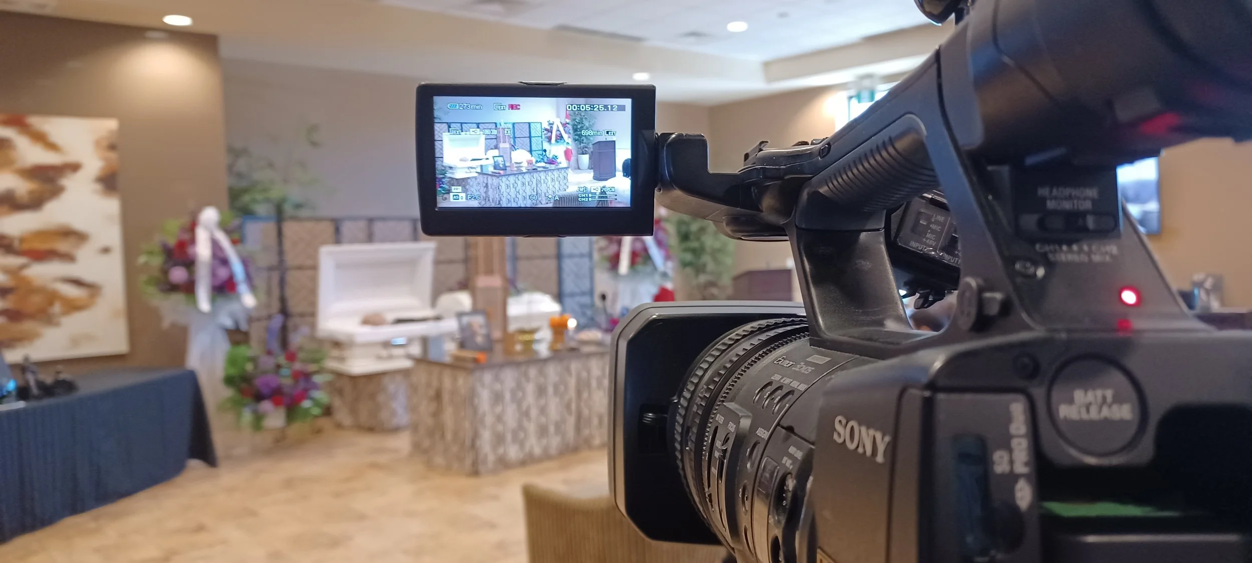 Camera filming a scene with a display screen showing a celebration set with a casket, flowers, and decorations in an indoor venue