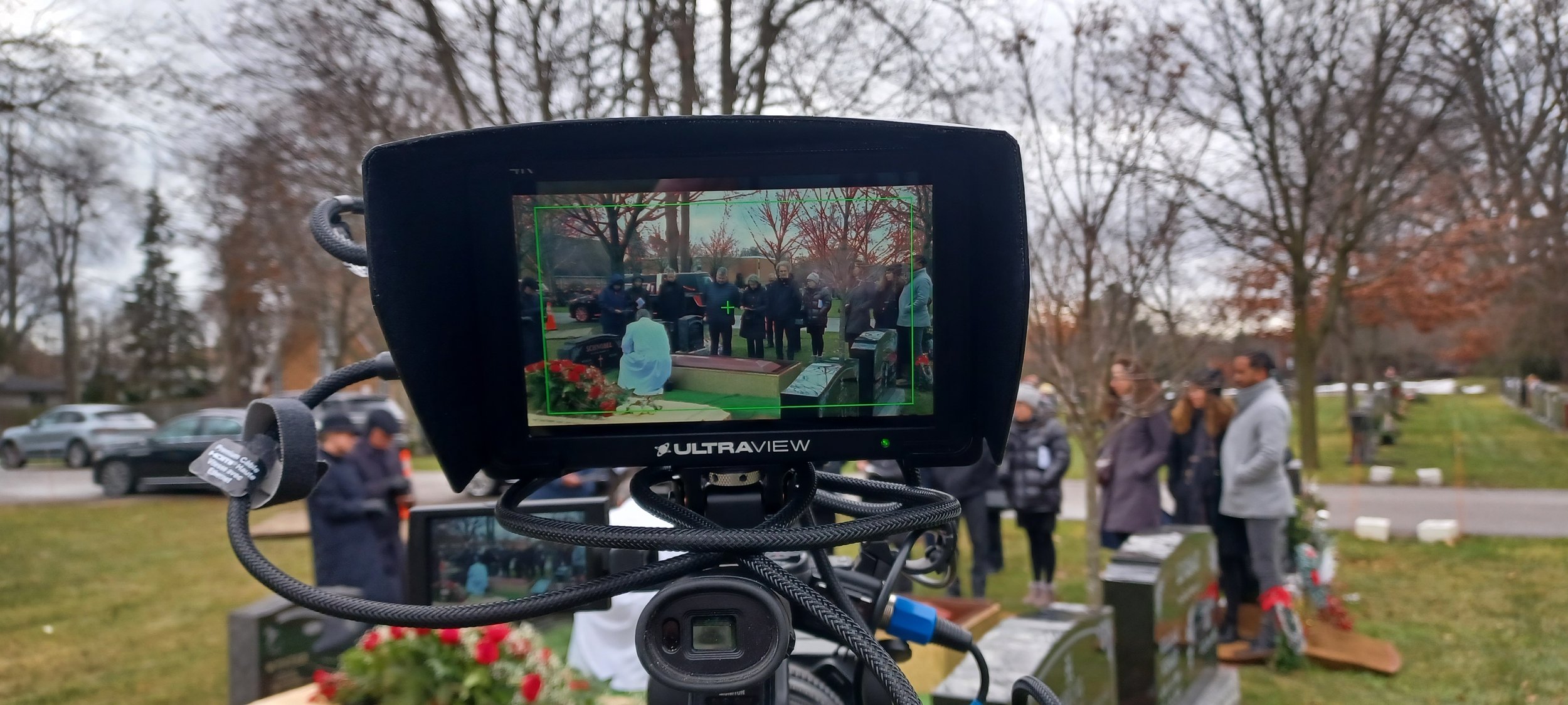 Camera filming a funeral ceremony at a cemetery with mourners gathered around a casket and floral arrangements during overcast weather.