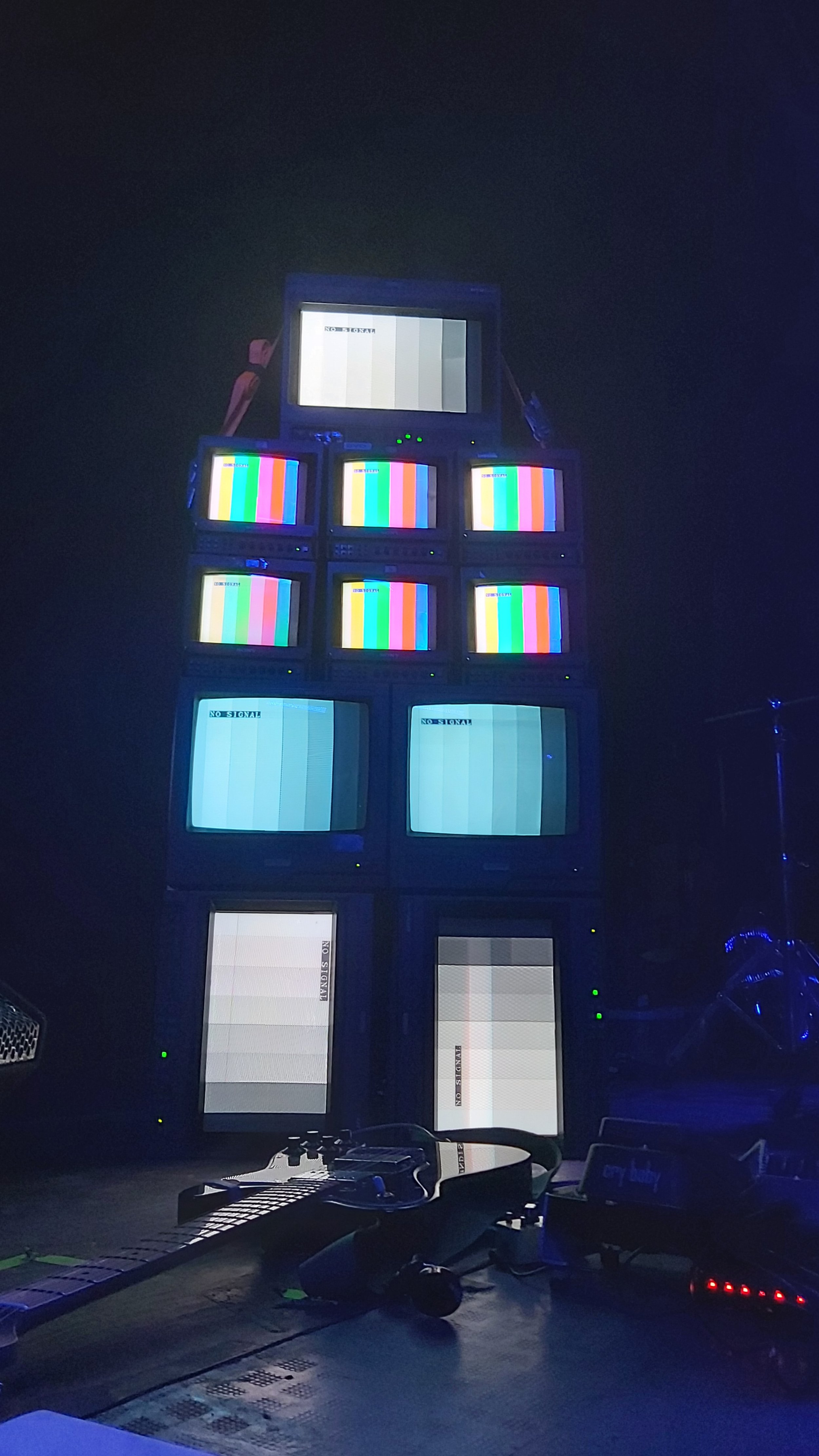 A dark room with multiple television screens stacked in a pyramid, displaying test pattern images and "No Signal" messages. There is a DJ setup with a turntable, mixer, and some electronic equipment on the floor in front of the screens.
