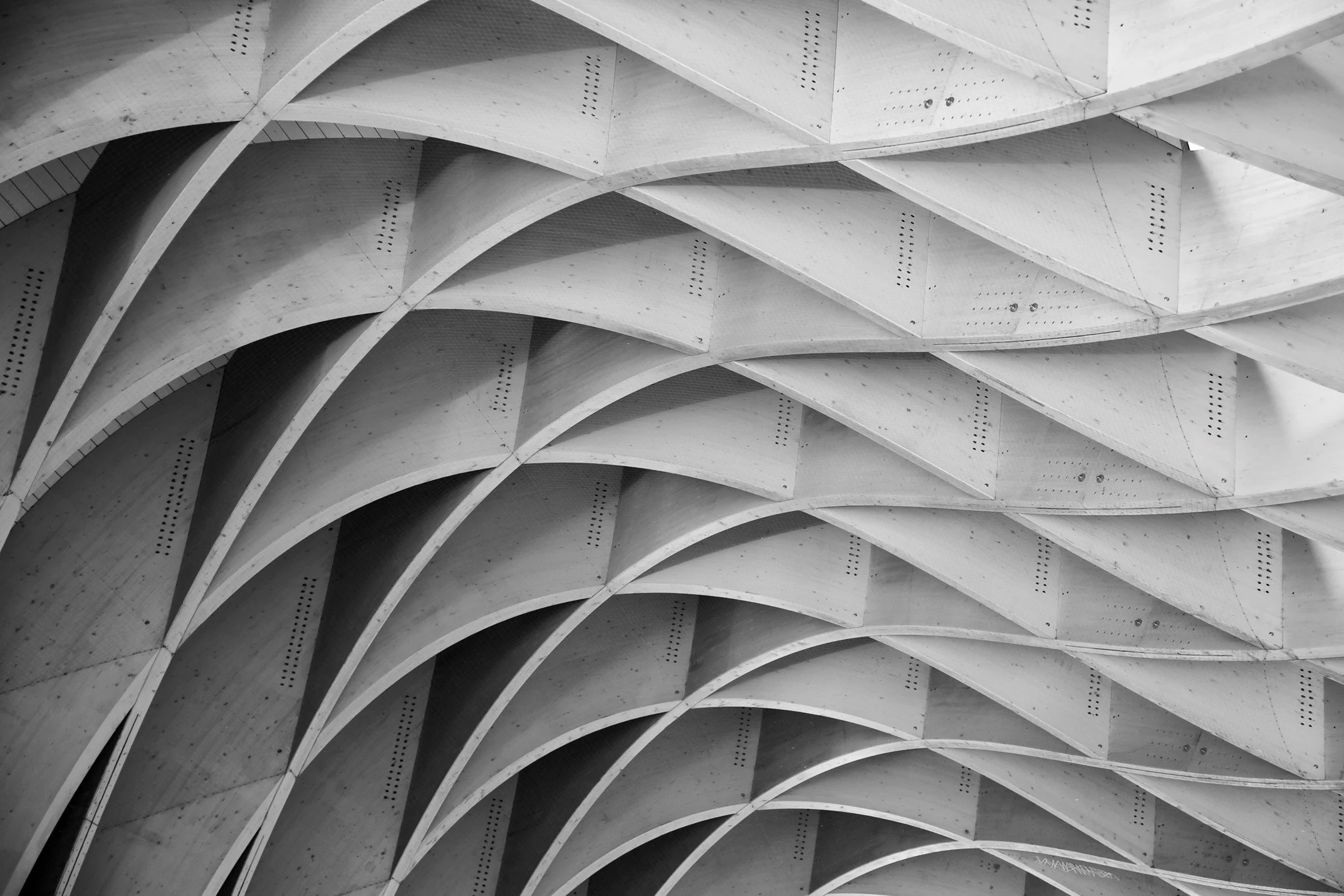 Close-up view of a modern architectural ceiling with repetitive curved, layered wooden or concrete beams, creating an intricate pattern.
