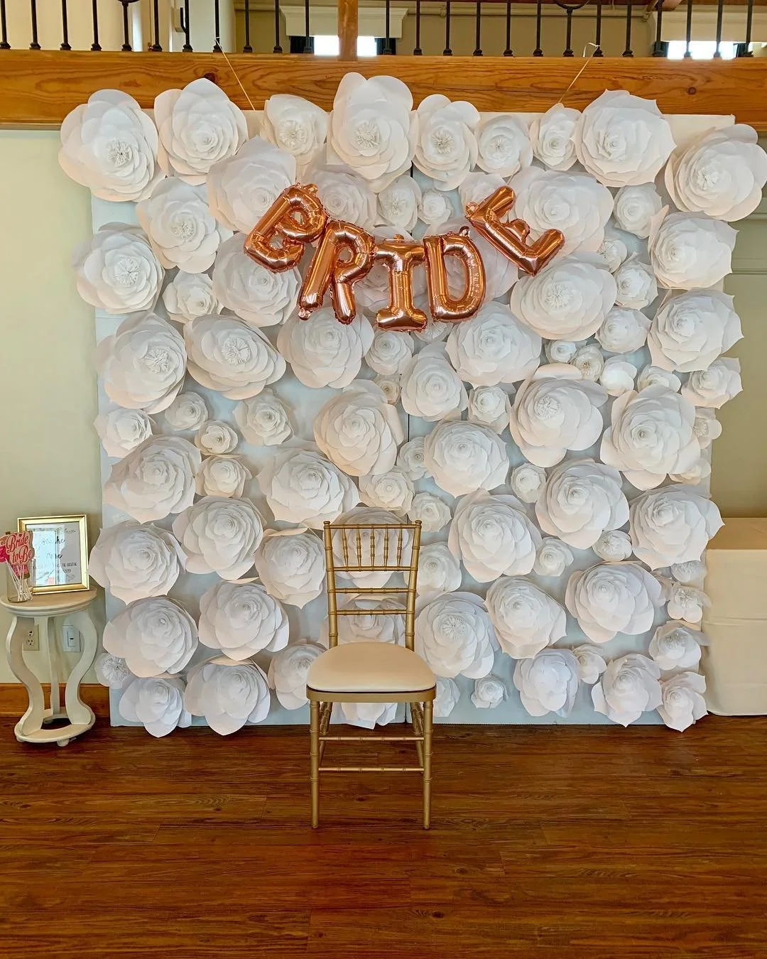 White floral backdrop with large paper flowers, rose gold balloon spelling "BRIDE", gold chair in front, small table with framed notes on the left.