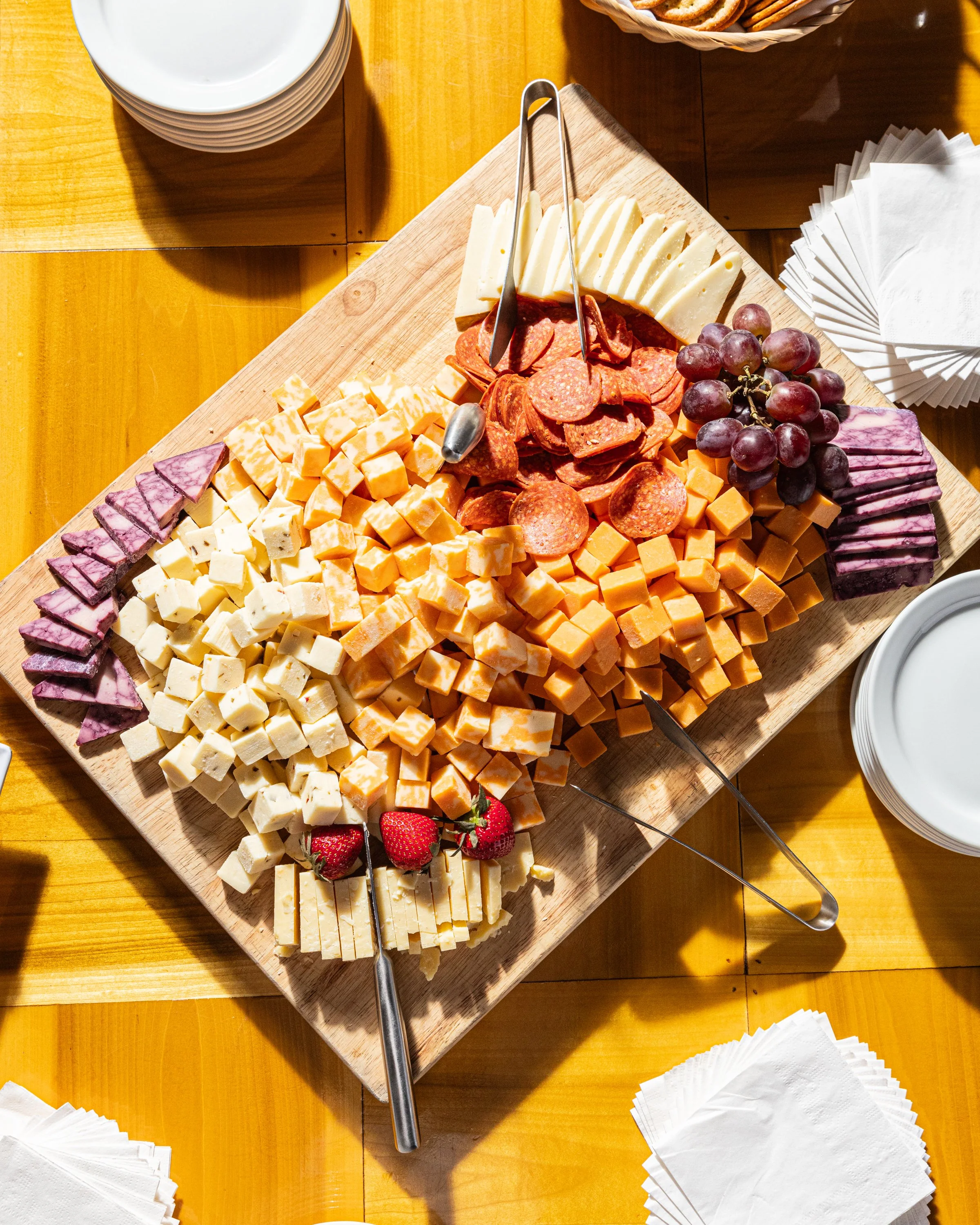 A cheese and meat platter featuring sliced cheese, salami, grapes, and strawberries arranged on a wooden cutting board, with plates, napkins, and a basket of bread visible in the background.