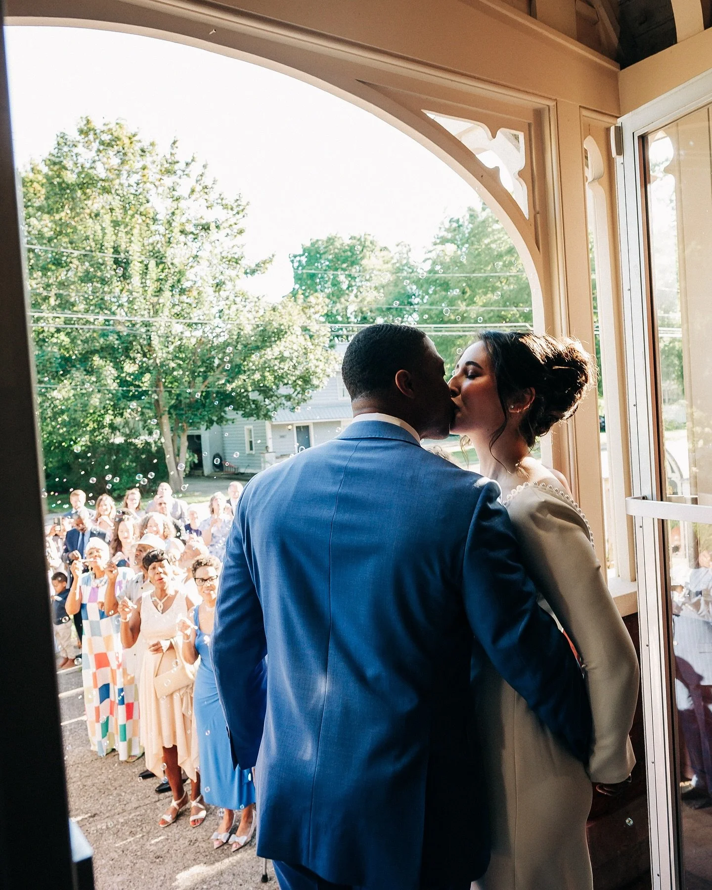 A bride and groom kissing inside a building, with guests outside watching and taking photos.