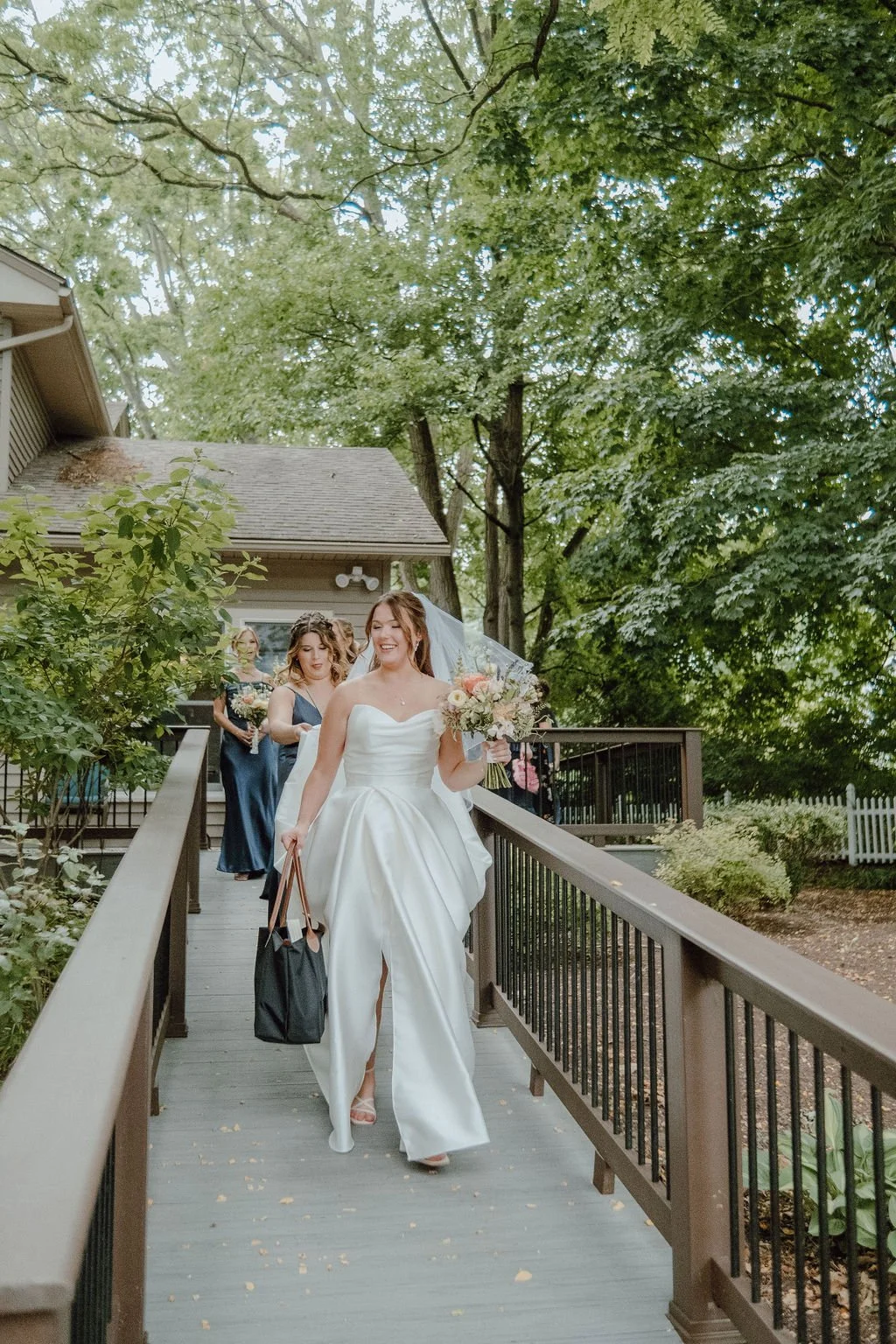 A bride in a white wedding dress holding a bouquet, walking down a path with bridesmaids behind her, surrounded by trees and greenery.