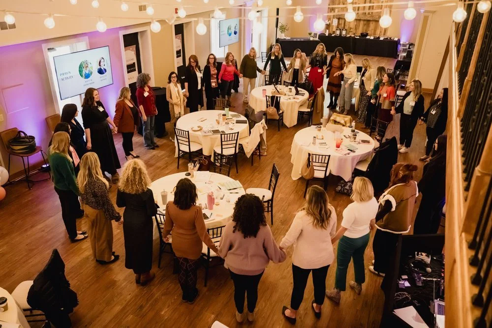 Women standing in a circle holding hands in a banquet hall with round tables, string lights, and presentation screens.