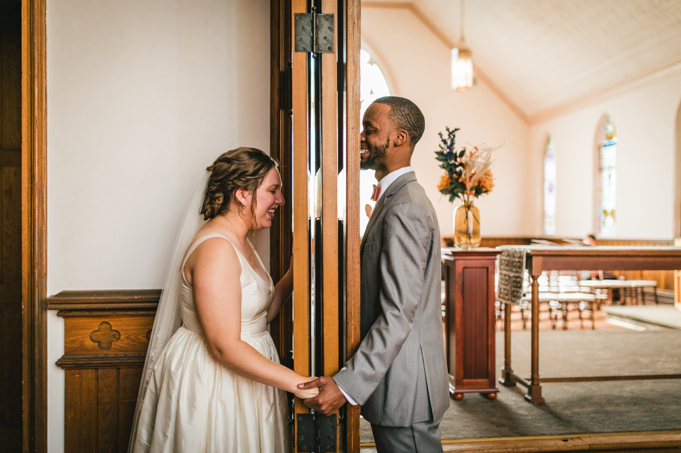 A bride and groom smiling and holding hands, separated by a door frame inside a church.