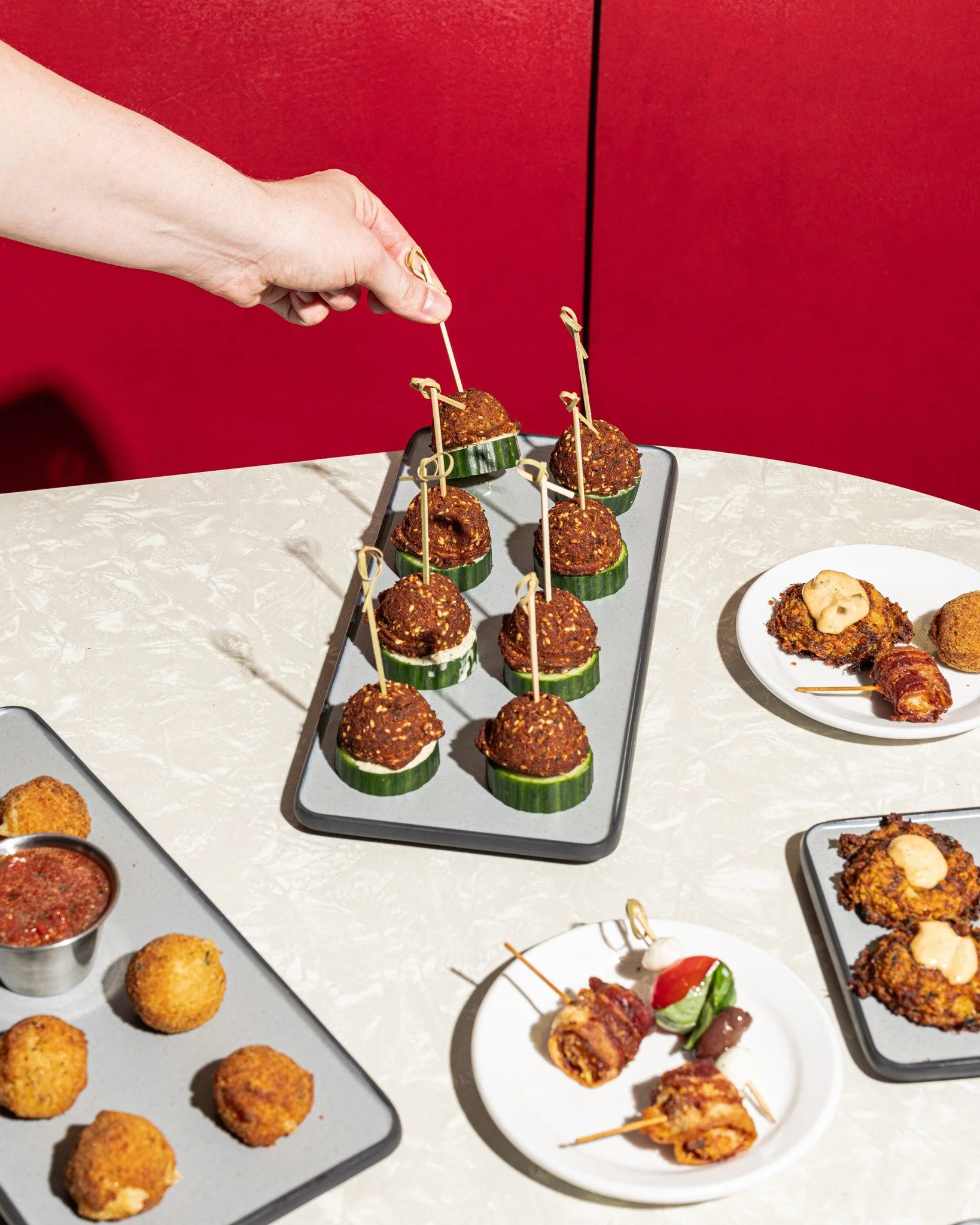 A hand picks up an appetizer on a black tray, with other similar appetizers on the tray and plates, including fried items and skewers, on a table with a red background.