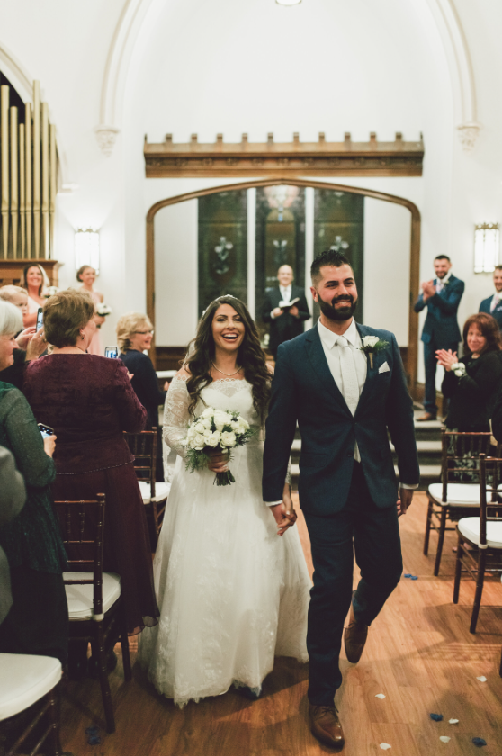 Bride and groom walking down the aisle, holding hands, smiling, with wedding guests clapping and celebrating in an elegant indoor venue.