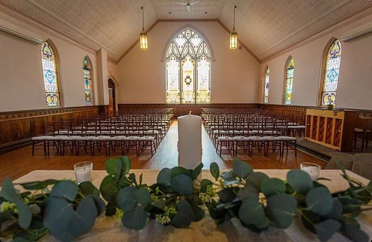 Interior of a church with wooden pews, stained glass windows, and a wooden altar decorated with green foliage and white candles.