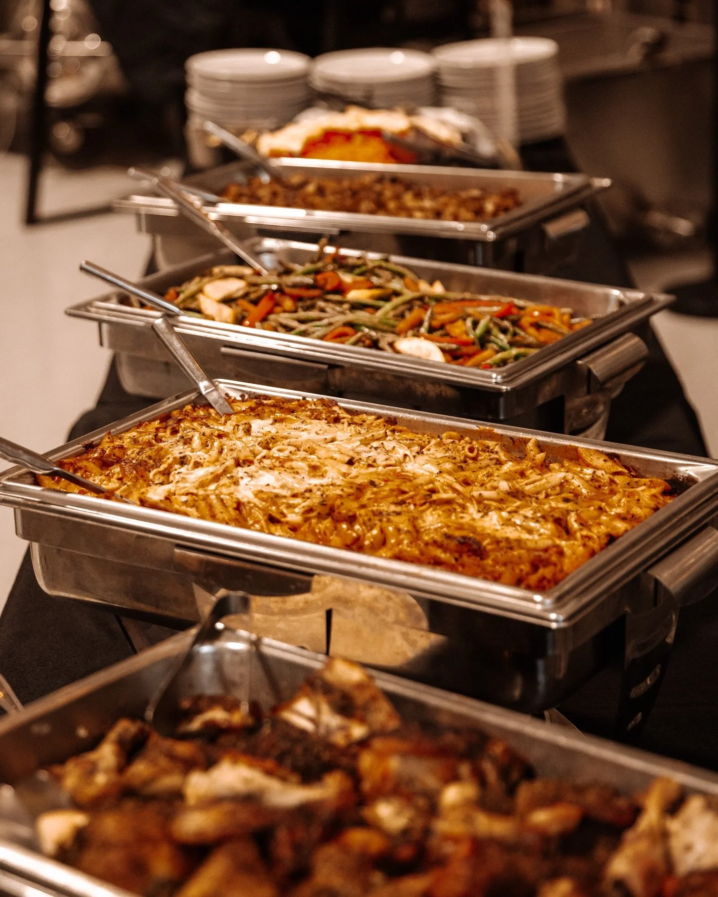 Buffet-style food with trays of baked pasta, sautéed vegetables, and other hot dishes on a serving table.