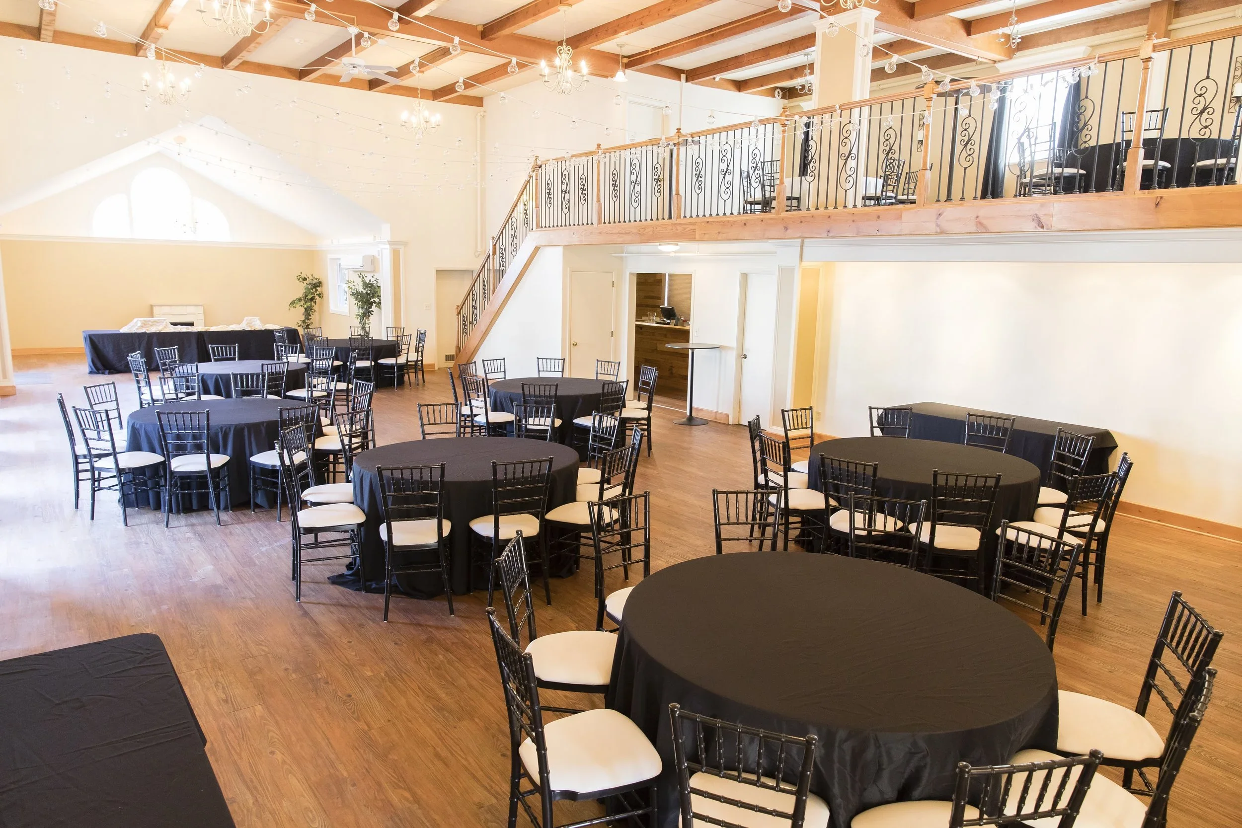 Event space with black round tables covered with black tablecloths, surrounded by white-cushioned black chairs, high ceilings with wooden beams, string lights, chandeliers, an upper balcony with chairs, and a staircase, illuminated by natural and artificial light.
