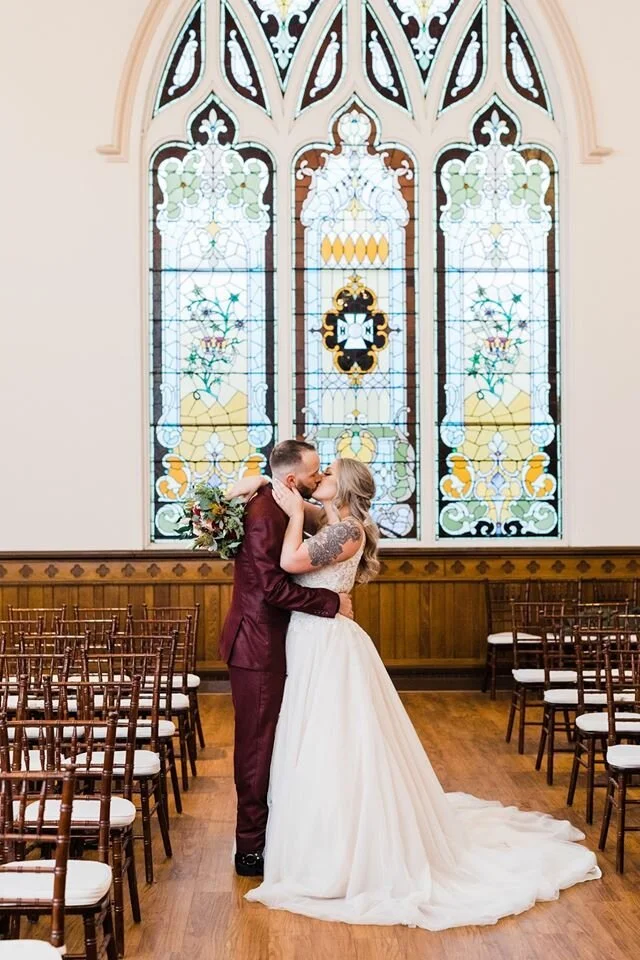 A bride and groom share a kiss inside a church with stained glass windows behind them, surrounded by empty chairs.