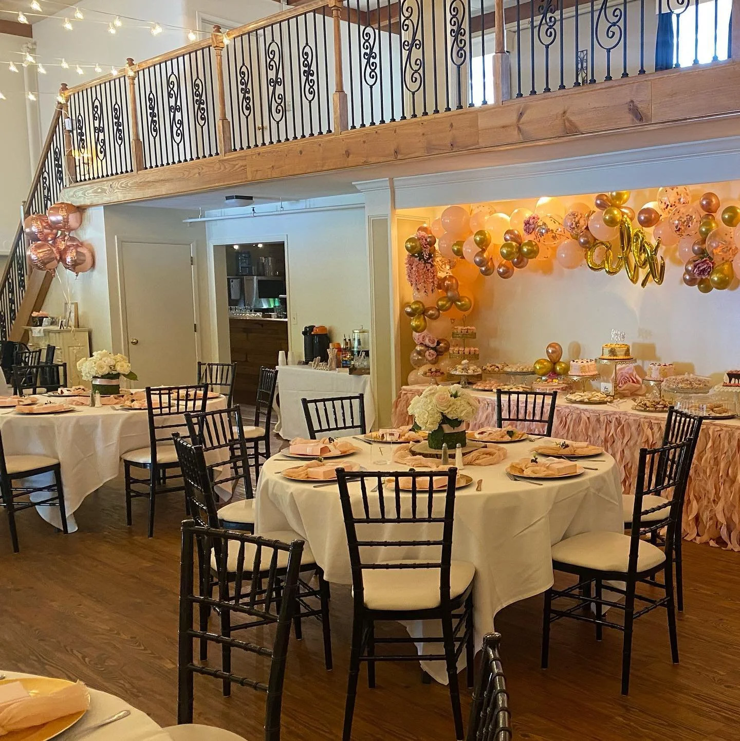 Decorated party room with round tables covered in white tablecloths, black chairs, floral centerpieces, and a dessert table with balloons and a "baby" sign for a celebration.