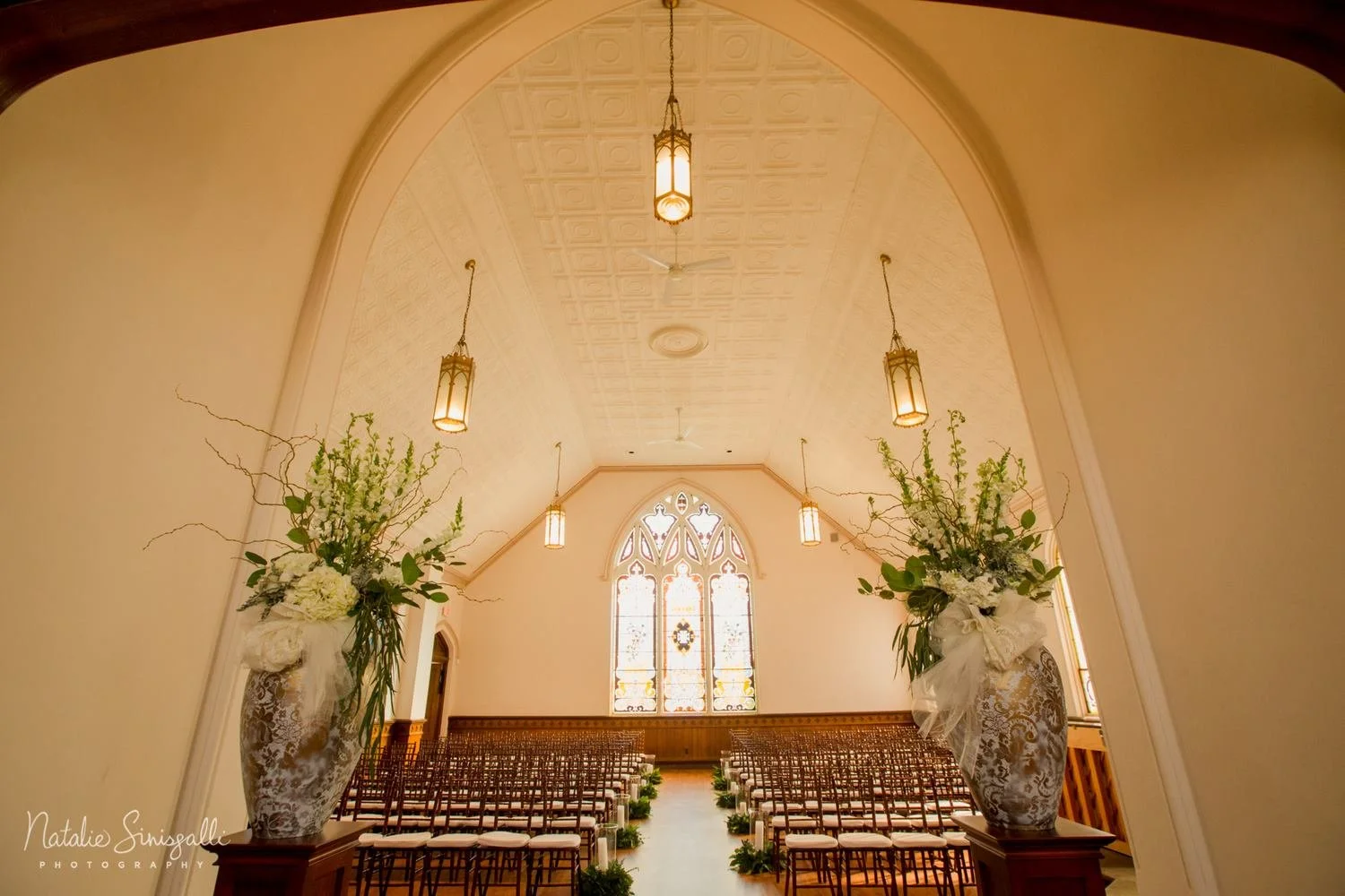 Interior of a church decorated for a wedding with large floral arrangements in vases and rows of chairs facing a stained glass window