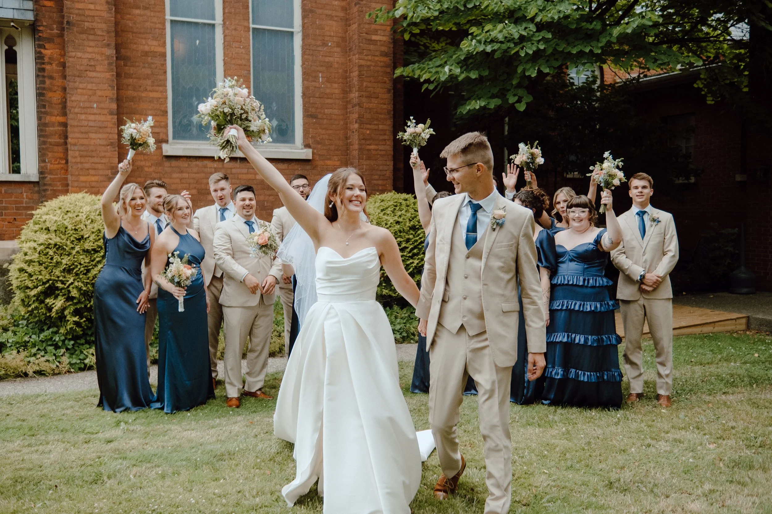 A bride and groom walking outdoors during their wedding celebration, surrounded by bridesmaids and groomsmen holding bouquets, with a brick building and greenery in the background.