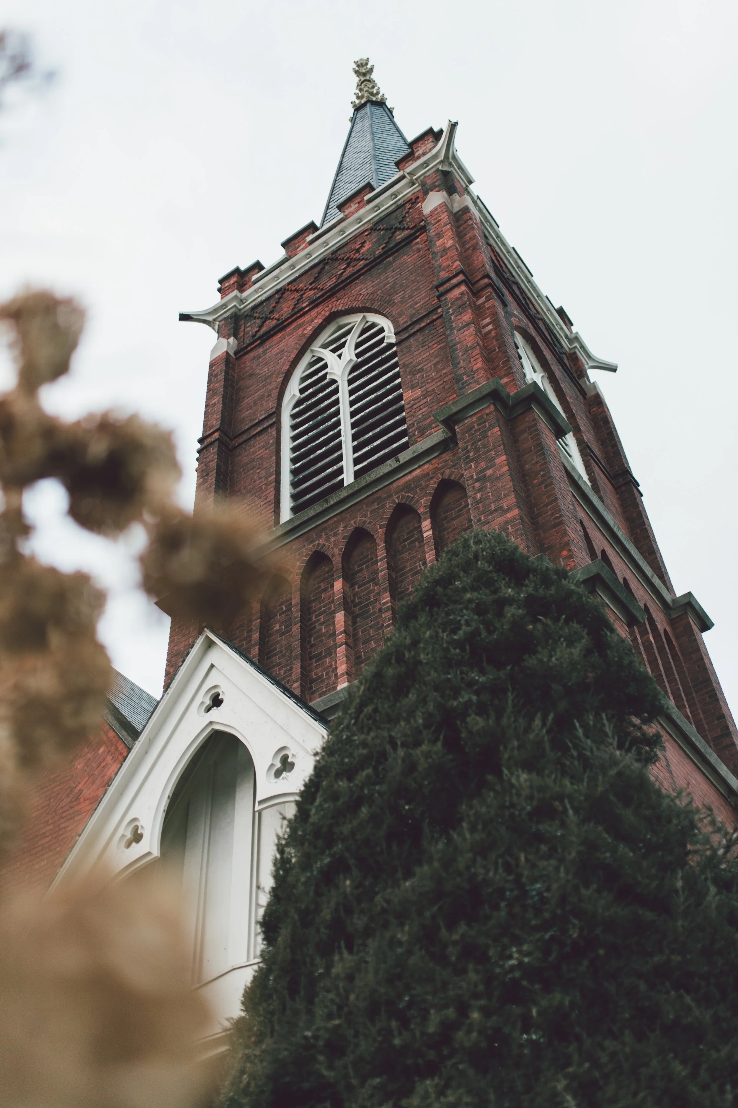 A tall, red brick church steeple with arched white window frames and a decorative shaped entrance, surrounded by green trees, viewed from below against an overcast sky.